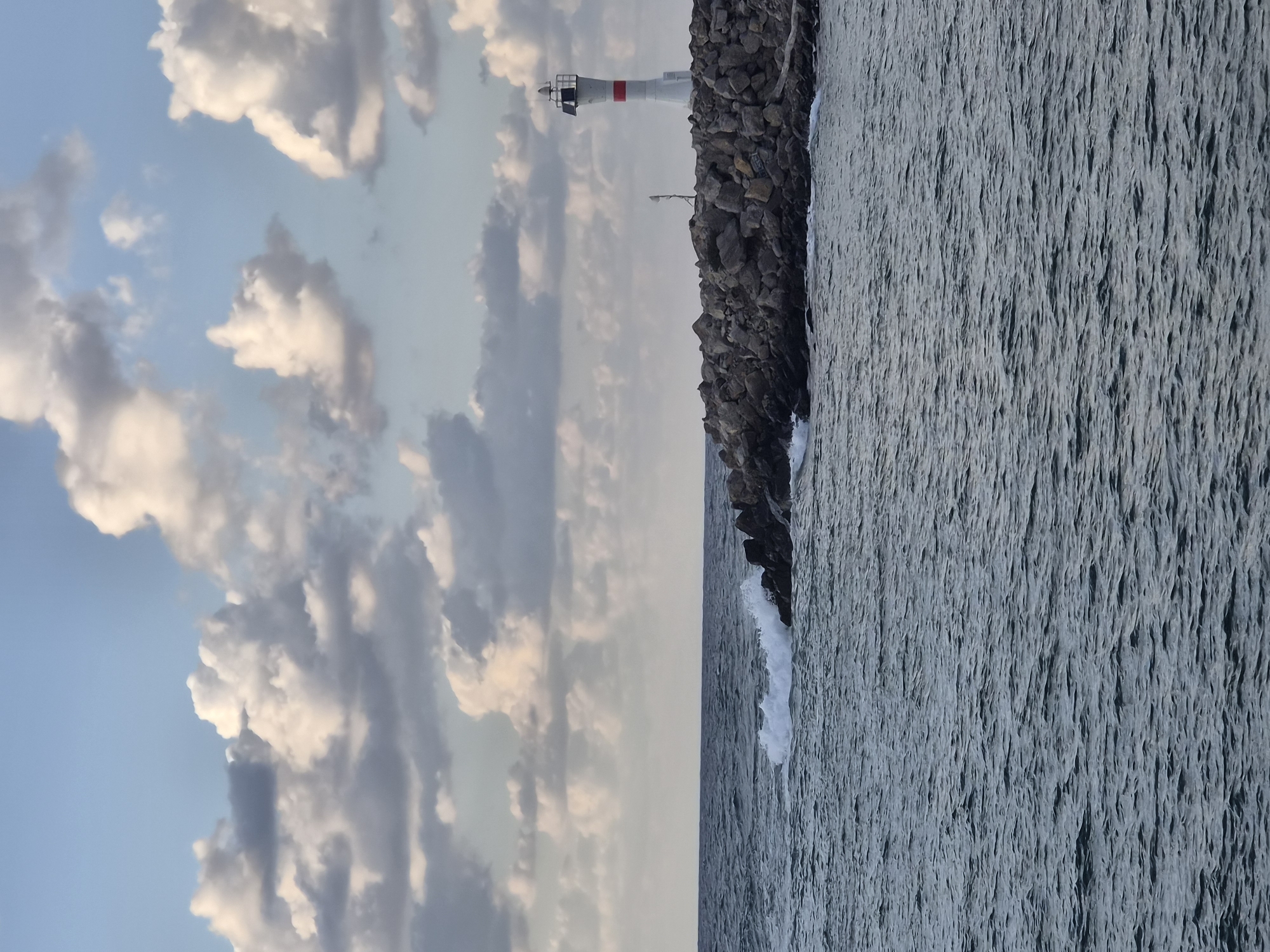 The image depicts a lighthouse situated on a rocky breakwater extending into the sea in Şile, Turkey. The lighthouse is white with a red band around its upper section, and it is topped with a navigational lamp. The breakwater is constructed from large, irregular grey stones. The sea in the foreground is dark blue with visible ripples and some white foam where waves are breaking against the rocks. The sky above is a pale blue, dotted with scattered white and grey clouds. The overall scene suggests a coastal environment under an overcast or partially cloudy sky.