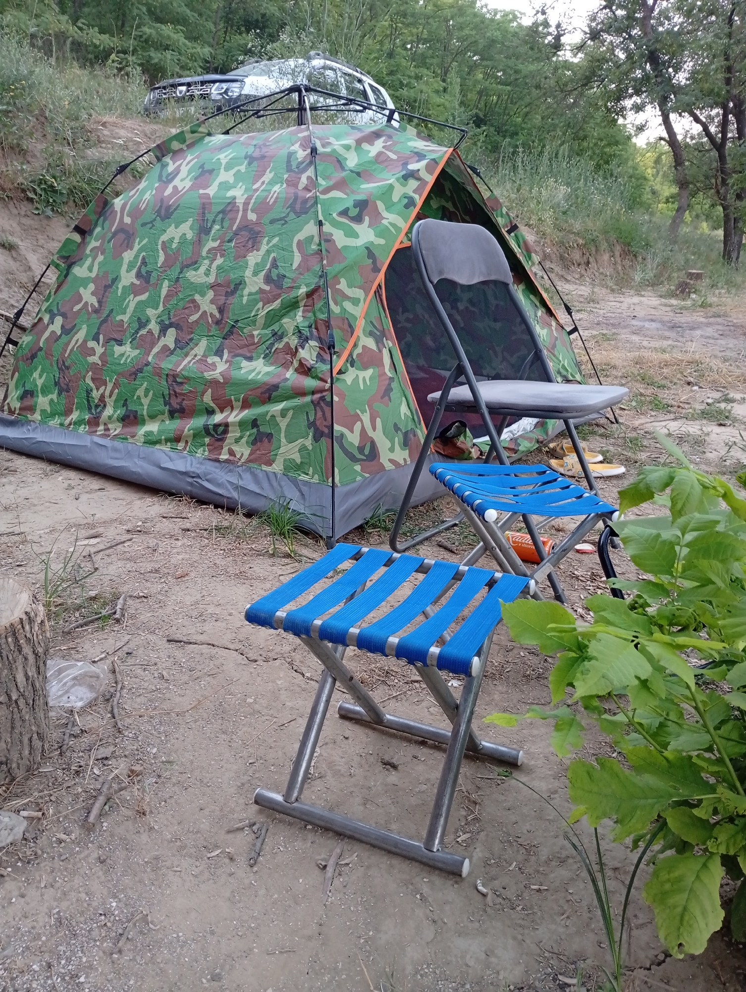 In Găleștii Noi, Moldova, a campsite is set up near a wooded area, with a camouflage-patterned tent as the central focus. In front of the tent are two blue folding stools and a larger grey folding chair. A silver SUV is parked on a higher ground in the background, partially obscured by vegetation. The ground is covered in dirt, small stones, and dried leaves, indicating an outdoor, possibly rustic, location. The lighting suggests it is daytime, possibly late afternoon or early morning, with soft light filtering through the trees. The scene appears to be untouched by human activity at the moment captured.