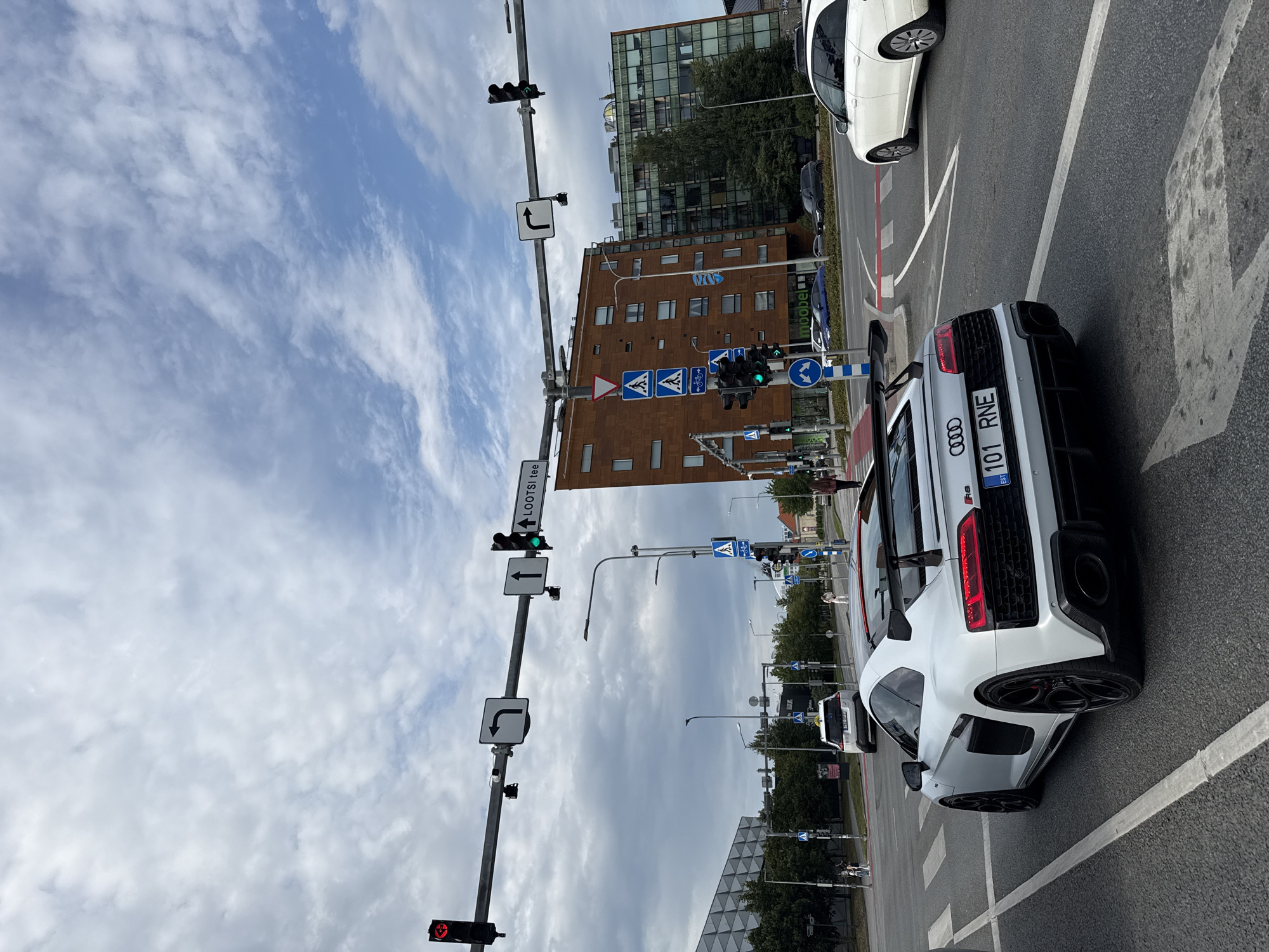 An Audi R8 sports car, license plate 101 RNE, is captured from the rear, angled slightly to the left, at a street intersection under a cloudy sky. The car is a light silver color and features a large rear spoiler and black wheels. In the background, a white station wagon is visible turning right, and further back, several buildings of varying architectural styles stand. Traffic lights and street signs are present at the intersection, with one sign indicating "LOOTSI tee" with an upward arrow and a right-turn arrow, suggesting this is a street in Tallinn, Estonia. The road surface is asphalt with visible white lane markings and a pedestrian crossing. Several pedestrians are sparsely present in the scene, including one on the far right of the frame walking away from the camera. The overall environment appears to be an urban setting in daylight.