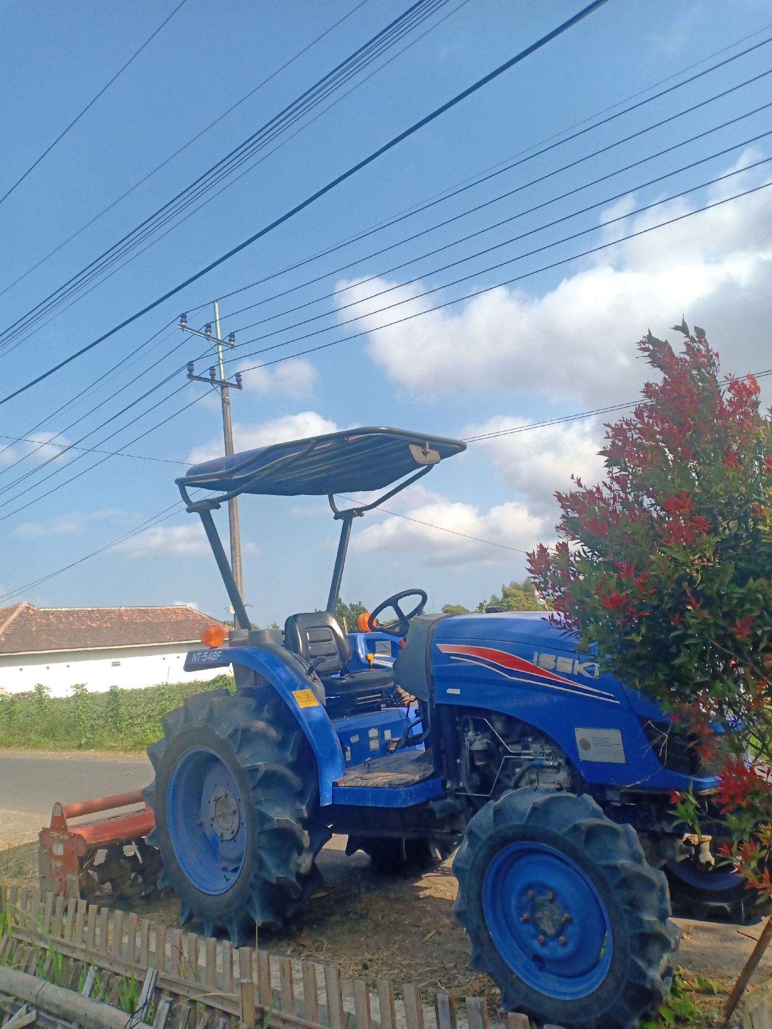 An outdoor, eye-level, medium shot captures a blue tractor with a black canopy, positioned on a dirt path next to a paved road. The tractor is the primary subject, showcasing its large, ribbed tires and the "ISEKI" logo on its side. A red tilling attachment is connected to the rear of the tractor.
In the background, a white building with a red-tiled roof is visible, partially obscured by green foliage. A wooden fence runs along the foreground, separating the tractor from the viewer. Overhead, a utility pole with multiple wires stretches across the bright blue sky, which is dotted with fluffy white clouds. To the right of the tractor, a tree with vibrant red leaves adds a splash of color to the scene. The overall environment suggests a rural or suburban setting.