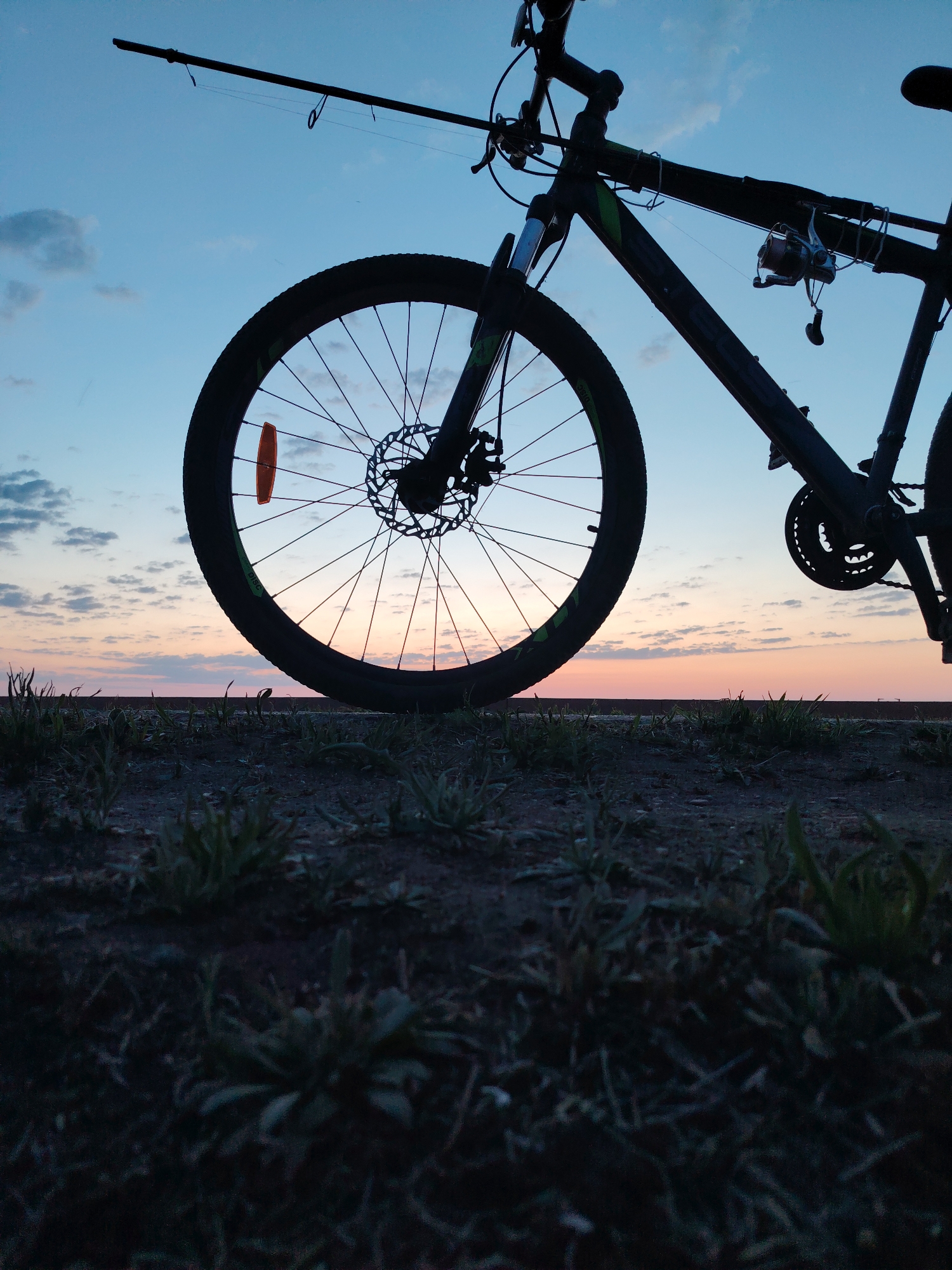 The image depicts a bicycle with a fishing rod attached to it, silhouetted against a sunset or sunrise sky. The bicycle is positioned on a concrete surface with vegetation growing in the foreground. The sky shows a gradient of colors ranging from blue to orange/pink. The location is identified as the city of Chaikovsky, Russia.