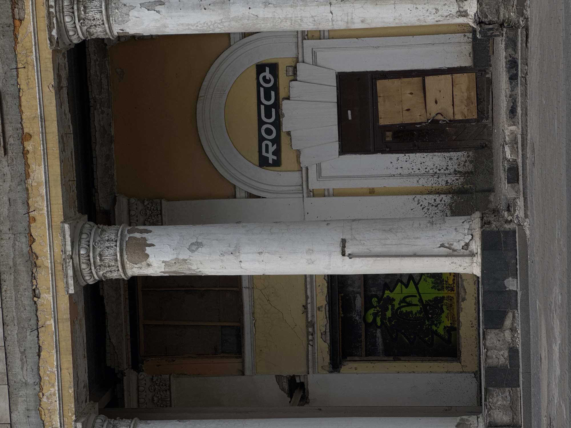 The image shows the exterior of a dilapidated building with classical architectural elements. Large, fluted columns, weathered and peeling, flank the entrance. Above the entrance, a dark sign with white lettering reads "ROCCO". The facade is painted a faded yellow, with visible cracks and water stains. To the left of the entrance, a window is boarded up and covered in bright green graffiti. The entrance itself is a dark wooden door, partially covered with plywood. The building appears to be in a state of disrepair, with debris and crumbling plaster visible on the ground and surrounding the entrance. The overall scene suggests an urban environment in a state of neglect or abandonment.