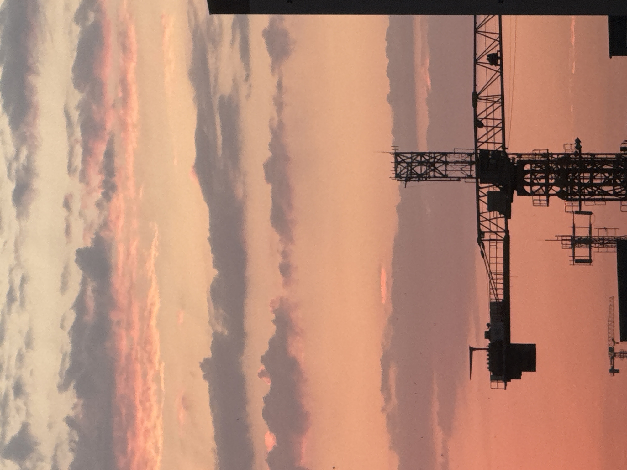 The image shows a construction site with several cranes silhouetted against a sunset sky. The sky is filled with clouds in shades of orange, pink, and gray. In the foreground, a large construction crane is prominently displayed, with its boom extending horizontally across the frame. Smaller cranes and structures are visible in the background, also silhouetted against the sky. The scene appears to be an urban environment, possibly Bishkek City, Kyrgyzstan, given the context provided. The lighting suggests it is either sunrise or sunset.