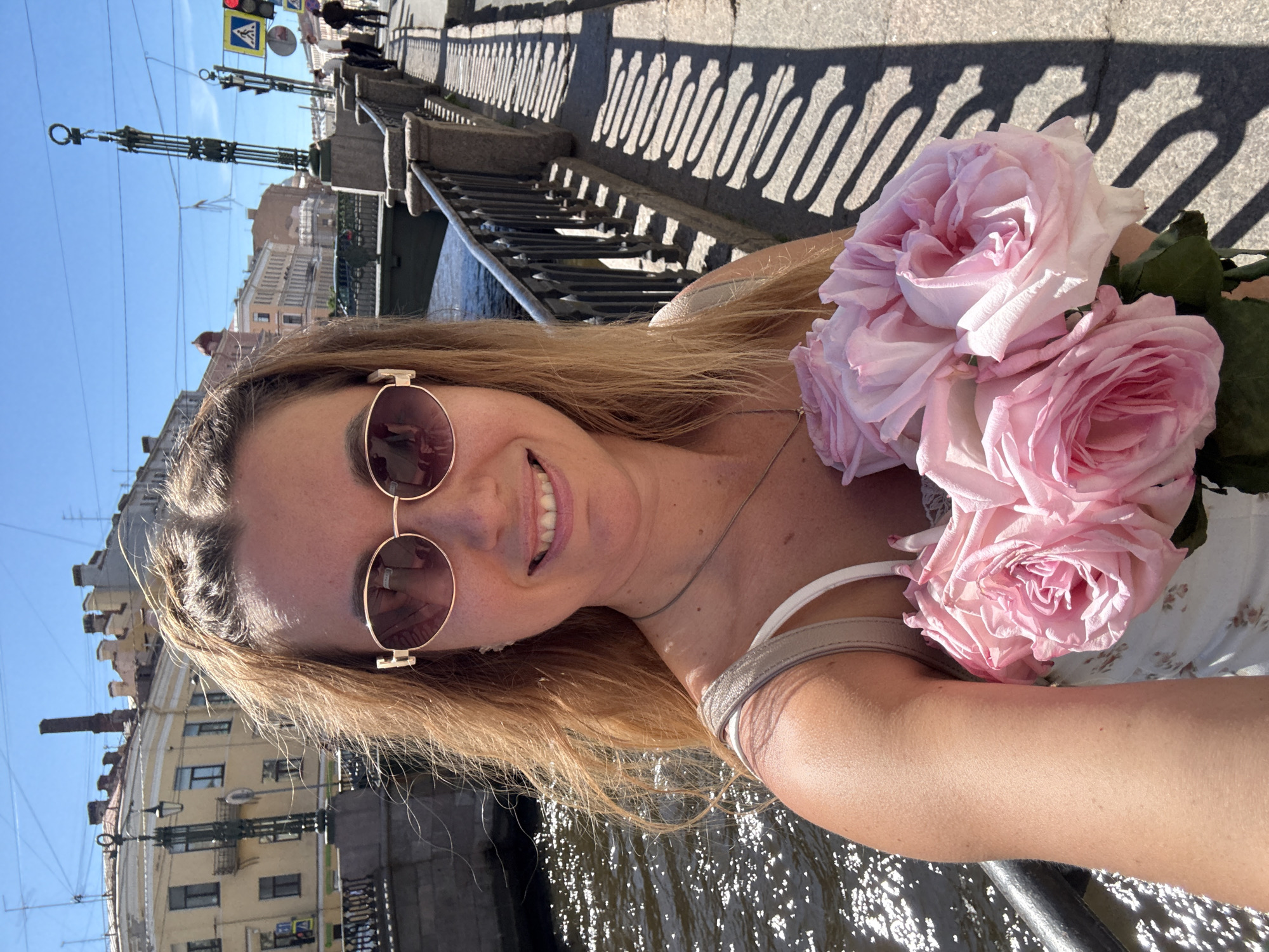 A young adult female is depicted in a close-up selfie, smiling directly at the camera. Her long, light brown hair is wind-swept. She wears oval-shaped sunglasses with gold-colored frames, reflecting elements of the surrounding urban environment, and a light-colored, possibly floral-patterned, strappy top with a thin silver necklace. She holds a bouquet of approximately six large, light pink roses, positioned in the lower-right foreground.
The background reveals an urban canal-side scene under a clear blue sky. To the left, a calm body of water, likely a canal, reflects bright sunlight. Lining the canal are multi-story buildings of classical European architecture, primarily in yellow and cream tones, featuring ornate windows, balconies, and chimneys visible on their roofs. A dark, decorative lamppost stands on the left bank.
To the right, a stone bridge with intricate dark metal railings crosses the canal. Beyond the bridge, on the sidewalk, strong, elongated shadows from the railing are cast, indicating bright sunlight and a high sun angle. A tall, decorative green structure, possibly a lamppost or a bridge mast, stands on the right side of the bridge. Further down the street, a red traffic light and a yellow triangular pedestrian crossing sign are visible, along with several indistinct pedestrians. The location is Unknown, Unknown.