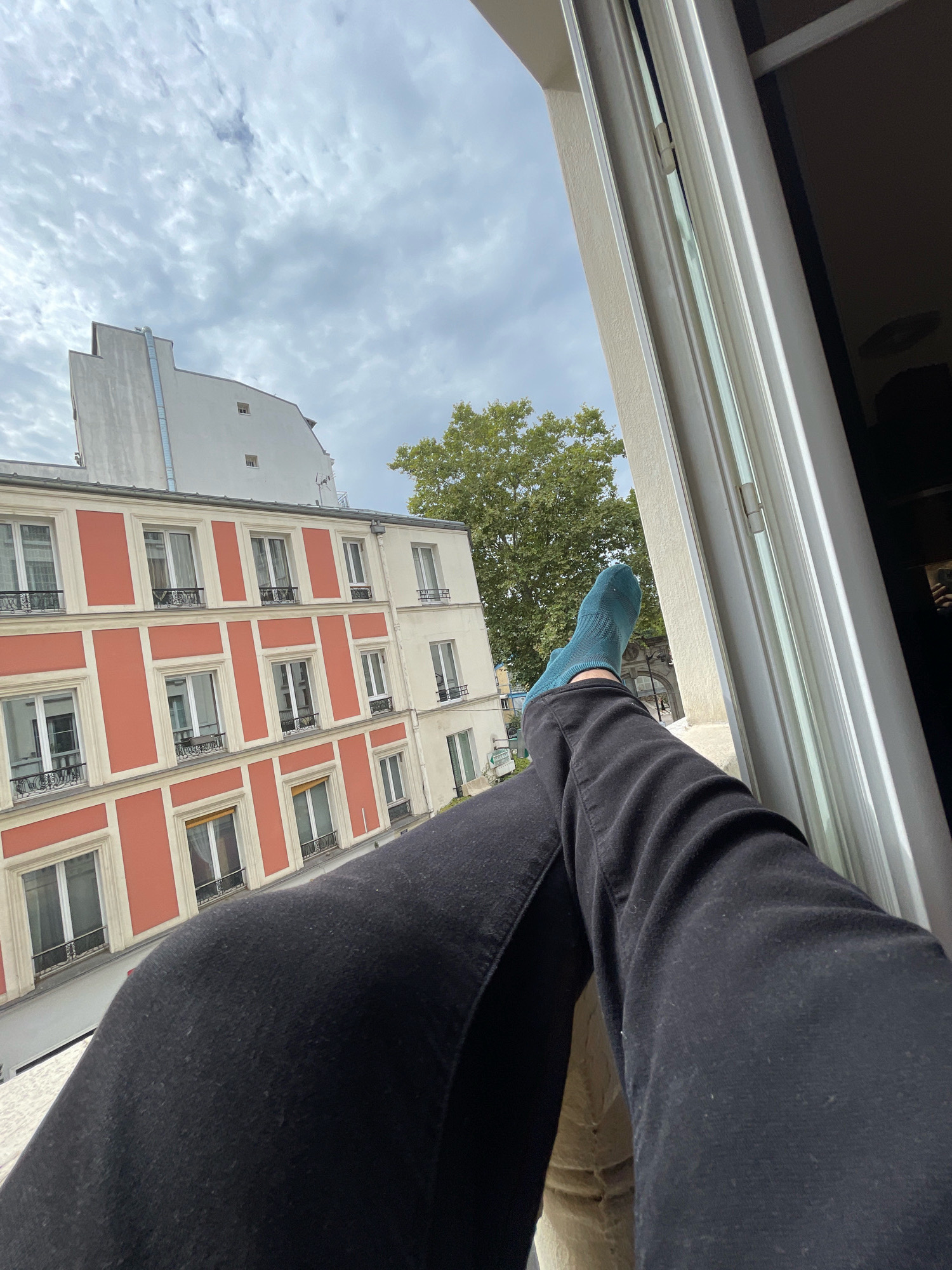 The image shows a pair of legs clad in black pants and teal socks, propped up on a windowsill. The feet are positioned such that the right foot, with the teal sock, is closer to the viewer and angled slightly upwards. The background reveals a view of Parisian architecture, characterized by multi-story buildings with distinctive red and cream facades, white window frames, and small balconies. The sky above is partly cloudy, with a few scattered clouds against a pale blue backdrop. A large green tree with dense foliage is visible to the right of the buildings. The perspective suggests the photo was taken from inside a building, looking outwards from a window. The overall scene evokes a relaxed, indoor moment with a view of the urban environment.