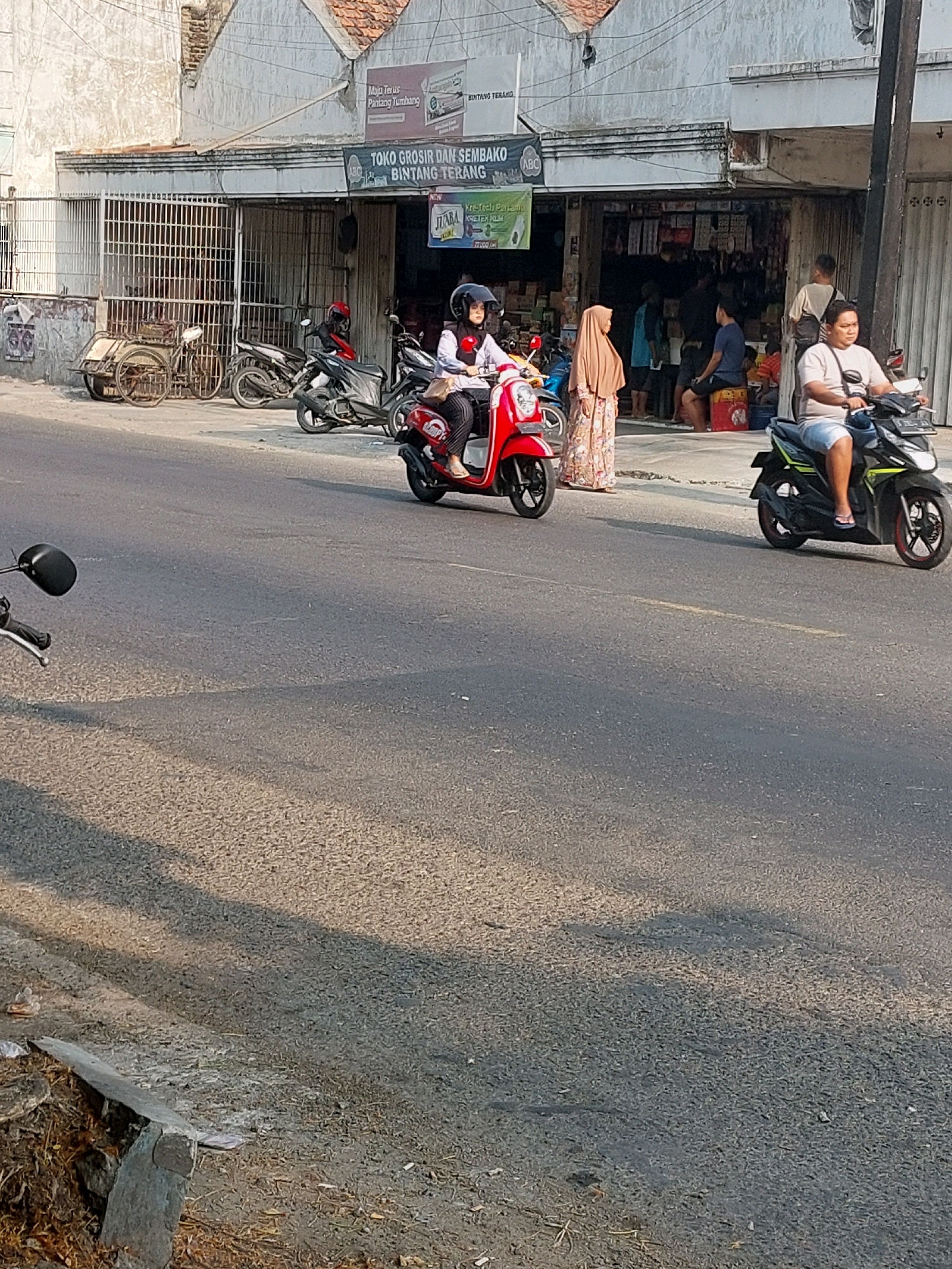 The image depicts a street scene in Sumuragung, Indonesia. In the background, a storefront with the signage "TOKO GROSIR DAN SEMBAKO BINTANG TERANG" is visible, indicating a wholesale grocery and provisions store. Several people are present, including a woman wearing a hijab and a white shirt riding a red scooter, and another woman in a patterned dress and hijab standing near the store. A man in a light-colored shirt and shorts is riding a dark-colored scooter on the right side of the street. Other motorcycles are parked or present on the street. The environment is a typical urban street with a paved road and sidewalks. The lighting suggests it is daytime.