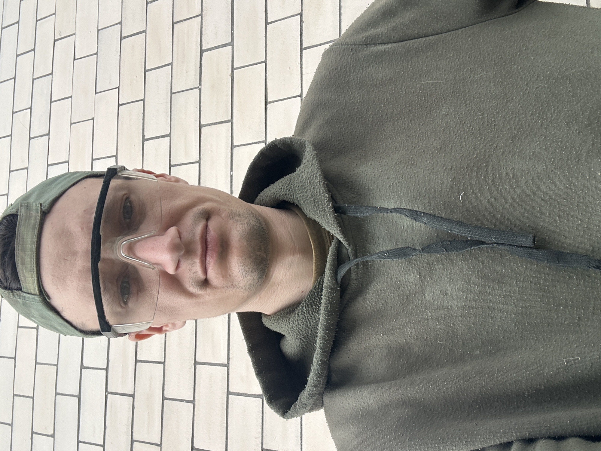 A medium shot selfie shows a man from the chest up. He is wearing a green baseball cap and clear safety glasses. His expression is neutral. He is wearing a green hooded sweatshirt over a tan t-shirt. The background is a wall made of off-white bricks. The image was taken outdoors in Makiv, Ukraine.