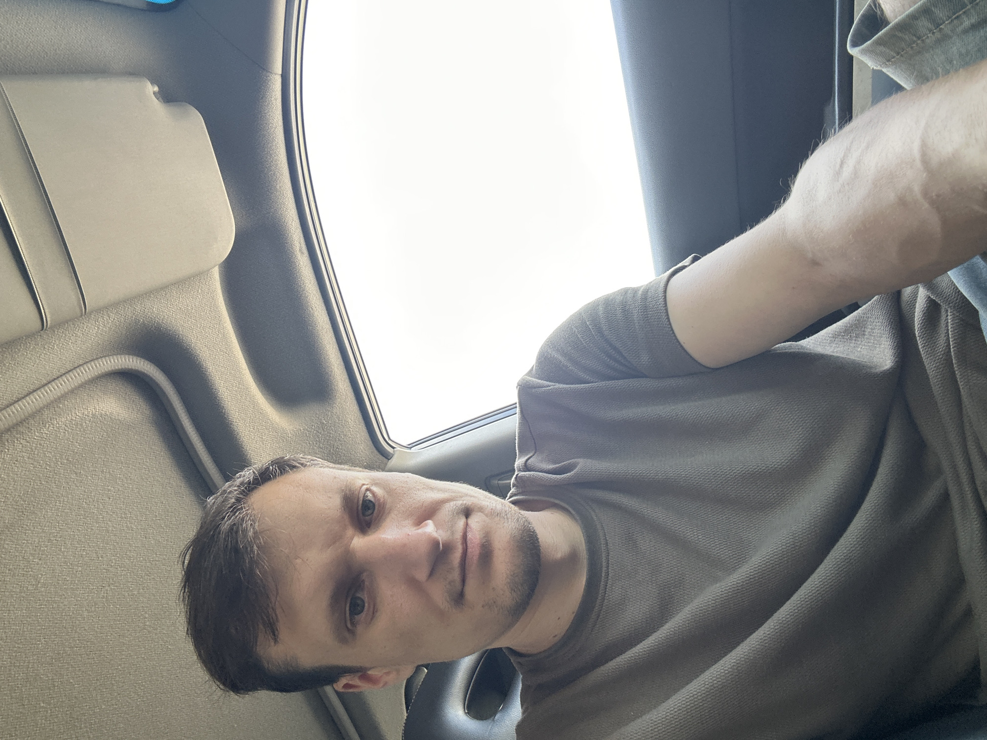 A man is seated inside a vehicle, with a sunroof visible overhead and a white, bright sky outside the window. The man is looking directly at the camera, his expression neutral. He is wearing a dark, short-sleeved t-shirt. The interior of the car appears to be a typical passenger vehicle. The scene is indoors, within the confines of the car. No specific location in Shatava, Ukraine can be identified from the visual information provided.