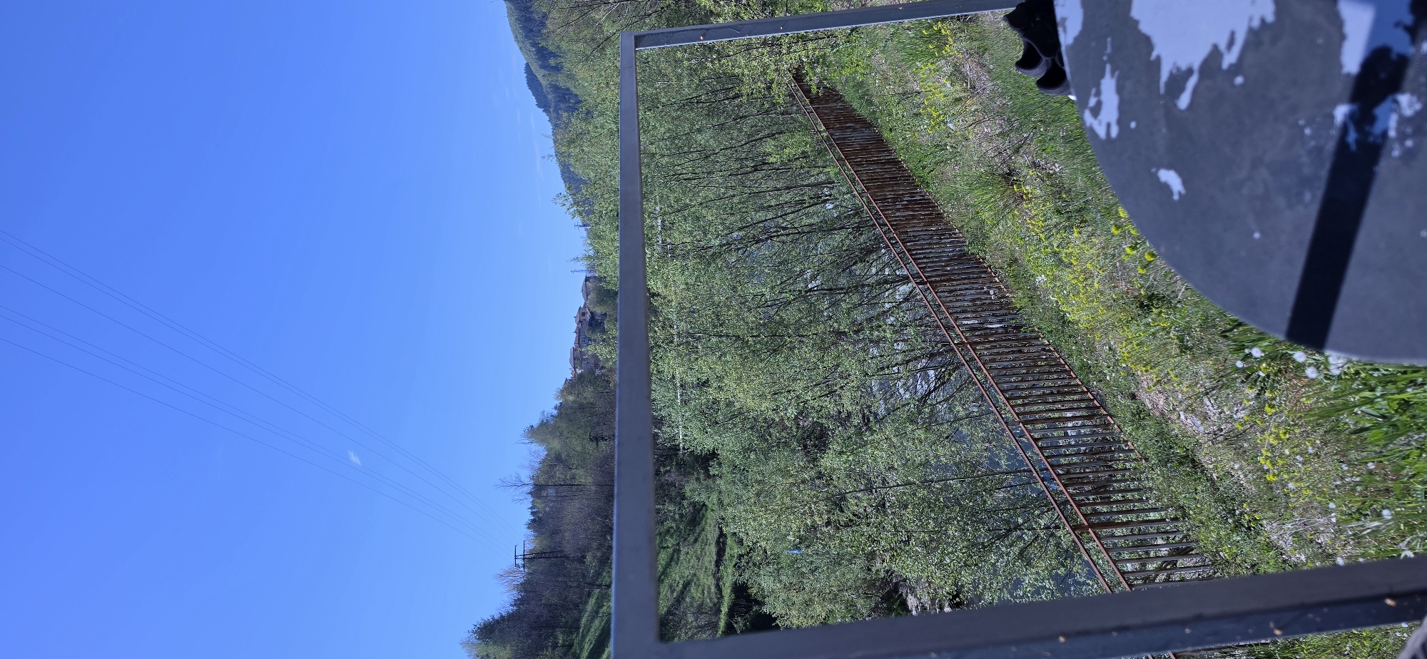 The image shows an outdoor scene. The viewpoint is from an elevated position, possibly a building or structure, looking down. The sky is clear and blue, taking up a significant portion of the frame on the left. Power lines are visible against the sky. On the right, there's a view through a metal frame, revealing a landscape with trees, vegetation, and a narrow stream or river flowing through the center. A rusted metal walkway or bridge spans the river. The vegetation includes trees with green foliage and grassy areas. The bottom part of the frame includes parts of the structure the photo was taken from, including the railing, along with some personal effects. The location is likely in a rural area near the city of Rudozem, Bulgaria, but there's no definitive proof from this image alone.