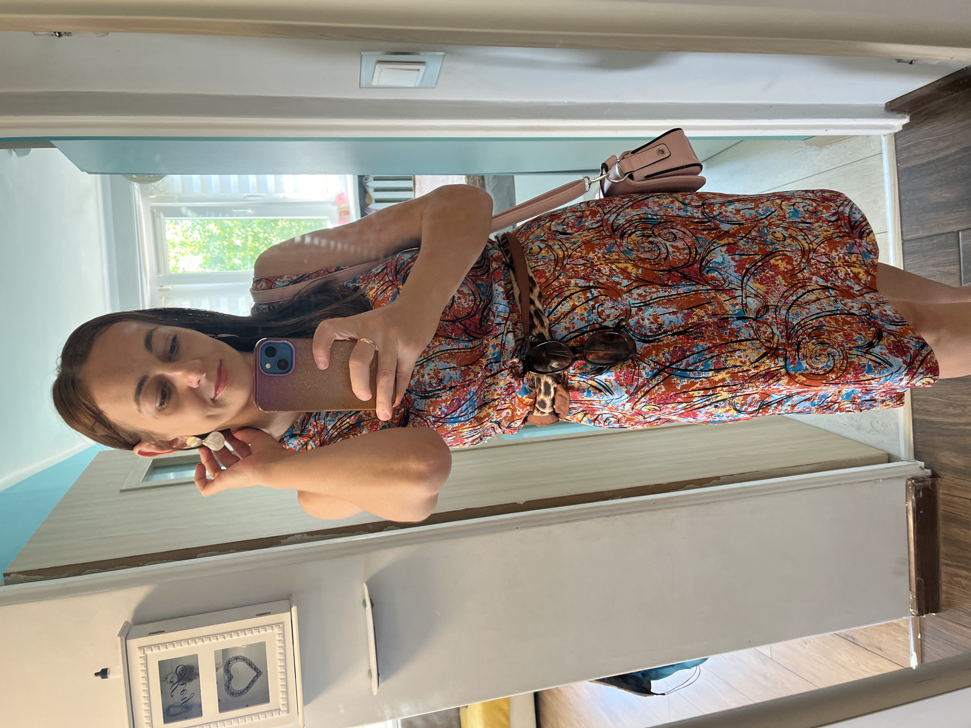 A woman is taking a mirror selfie in what appears to be a hallway. She is wearing a colorful, patterned dress with a leopard print belt and a light pink purse slung over her shoulder. Her hair is dark and pulled back, and she is wearing earrings and a ring. She is holding her phone up to take the photo. In the background, there is a window with trees visible outside, suggesting it might be daytime. There is also a framed picture on a shelf below the mirror. The setting is domestic, likely a home.