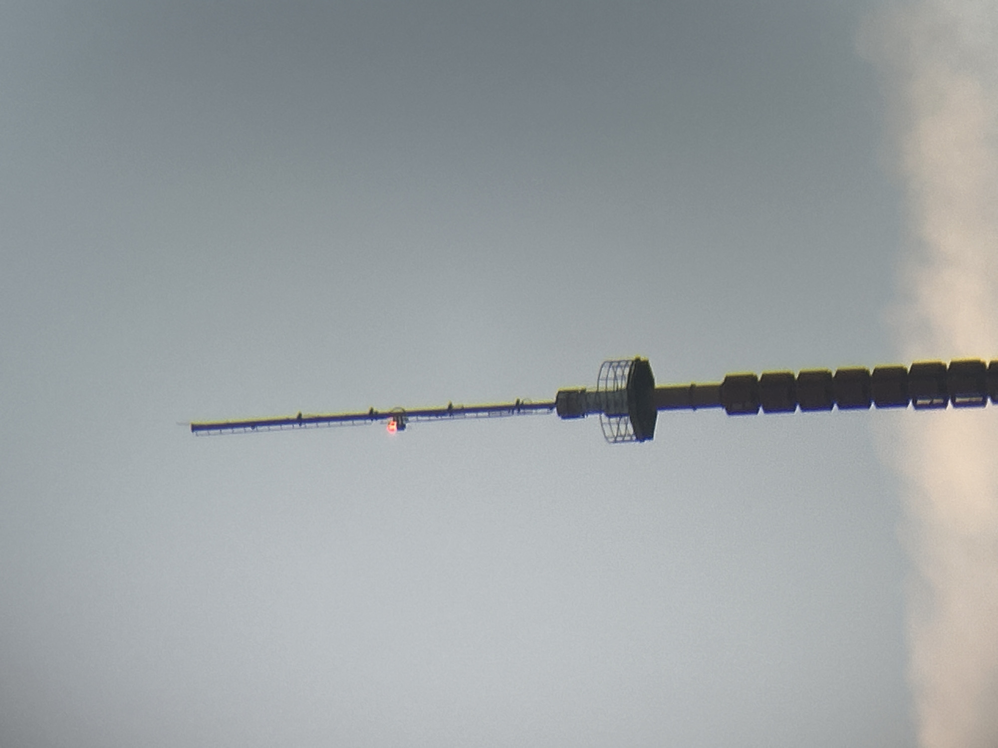 The image displays a horizontal segment of a tall, man-made structure against a largely overcast sky. This structure, likely part of a broadcast tower or mast, extends across the middle of the frame.
On the left side, the structure appears as a slender, linear beam. A distinctive, small, bright orange-red light is visible on this slender section, positioned roughly to the left of the image's center. Further to the right of this light, there is a cylindrical or disc-shaped component encased within a metallic, cage-like or lattice structure. The right portion of the structure consists of darker, rectangular, segmented blocks, appearing robust and uniformly arranged.
The background is an expansive, muted gray sky, indicating an overcast day, or potentially twilight hours. The sky lightens towards the bottom and left, while a brighter, more cloud-defined area is visible on the far right side of the frame. There is no visible human activity or discernible text within the image. Given the context of Tallinn, Estonia, this structure is consistent with parts of the Tallinn TV Tower, with the orange-red light functioning as an aviation obstruction warning.