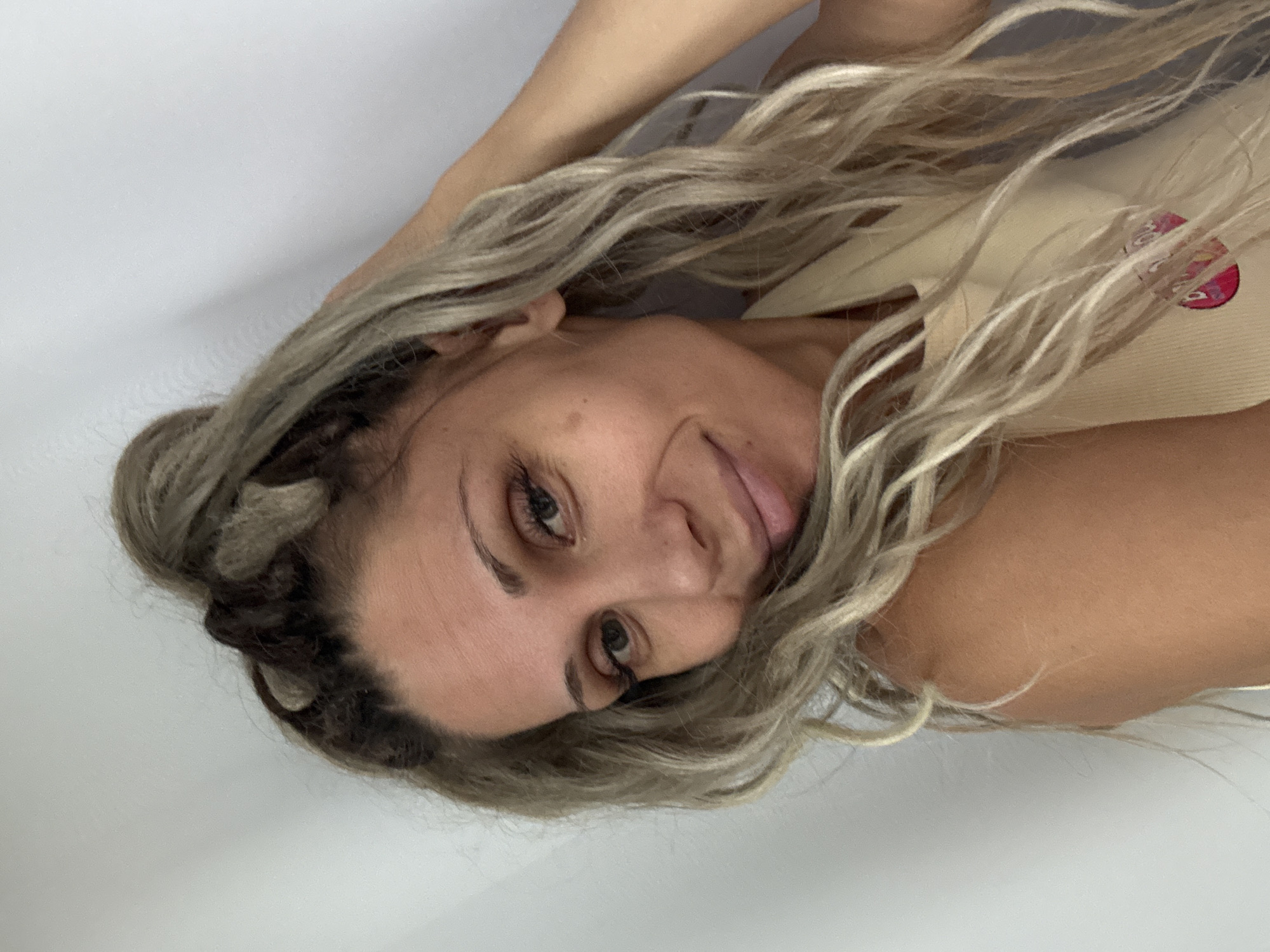 A medium close-up shot shows a fair-skinned woman with blonde hair styled in a messy bun on top of her head, with loose strands framing her face. Her hair is wavy and appears to be a mix of dark roots and lighter blonde ends. She is looking towards the camera with a slight smile, her head tilted and her right hand is touching her hair. Her makeup includes dark eyeliner and mascara, and her eyebrows are well-defined. She is wearing a light-colored tank top. The background is a plain, light-colored wall. There is no identifiable information about the location of Rusanovo, Russia.