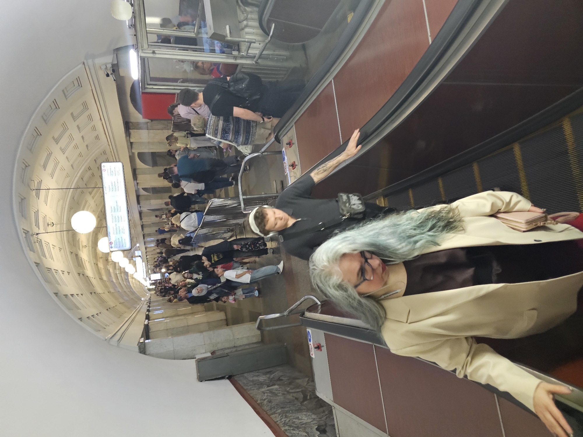 The image captures a bustling underground subway station, showcasing a long escalator system filled with commuters. In the foreground, standing on the right escalator, is a person with distinctive long, grey and blue hair, wearing glasses, a dark top, and a light beige blazer, holding a small pink wallet or purse. Behind them and slightly to the left is another individual with short, light-colored hair (possibly covered by a white cap or bandana), dressed in a dark t-shirt and carrying a black cross-body bag, with visible tattoos on their left arm.
Numerous other passengers are visible descending the escalators, creating a crowd that extends to the platform below. The station features an impressive vaulted ceiling adorned with intricate white, geometric paneling and illuminated by both circular dome lights and elongated fluorescent fixtures. Along the escalator shaft, the walls exhibit patterned designs, and the floor at the bottom appears to be made of patterned stone or tile. A red emergency stop button with a white hand symbol is present on the escalator's side panel.
A long, vertical, illuminated information sign hangs from the ceiling, displaying text in Cyrillic, notably "К пригородным поездам" (To suburban trains), indicating the location is likely in a Russian-speaking country. The scene depicts a typical busy public transport environment, with people engaged in their daily commute. The station is brightly lit by artificial light, providing no indication of the time of day.