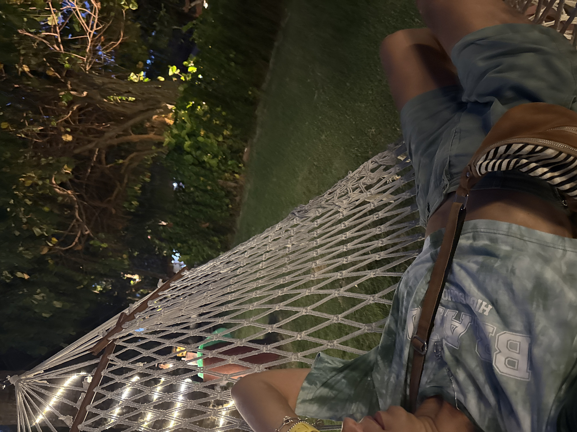 A person lies in a hammock at night in Kuta Selatan, Indonesia. They are wearing blue-grey shorts and a tie-dye t-shirt, with a brown bag slung over their shoulder. The hammock is strung between trees, adorned with small, warm-toned lights. A tree and green grass are behind the hammock.