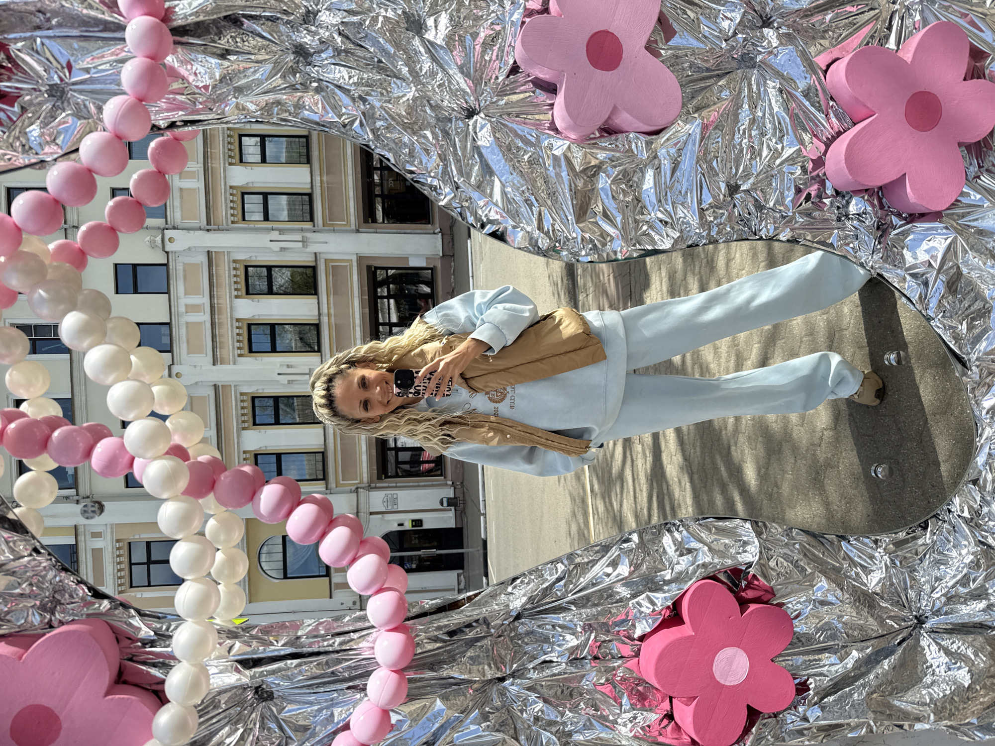 A woman is seen in a mirror taking a selfie. The mirror is on the ground surrounded by silver foil, pink flower decorations, and pink and white beads. The reflection in the mirror shows the woman wearing a light blue sweatsuit and a brown jacket, holding her phone. The background reflected in the mirror shows a building in the city of Unknown, Unknown.
