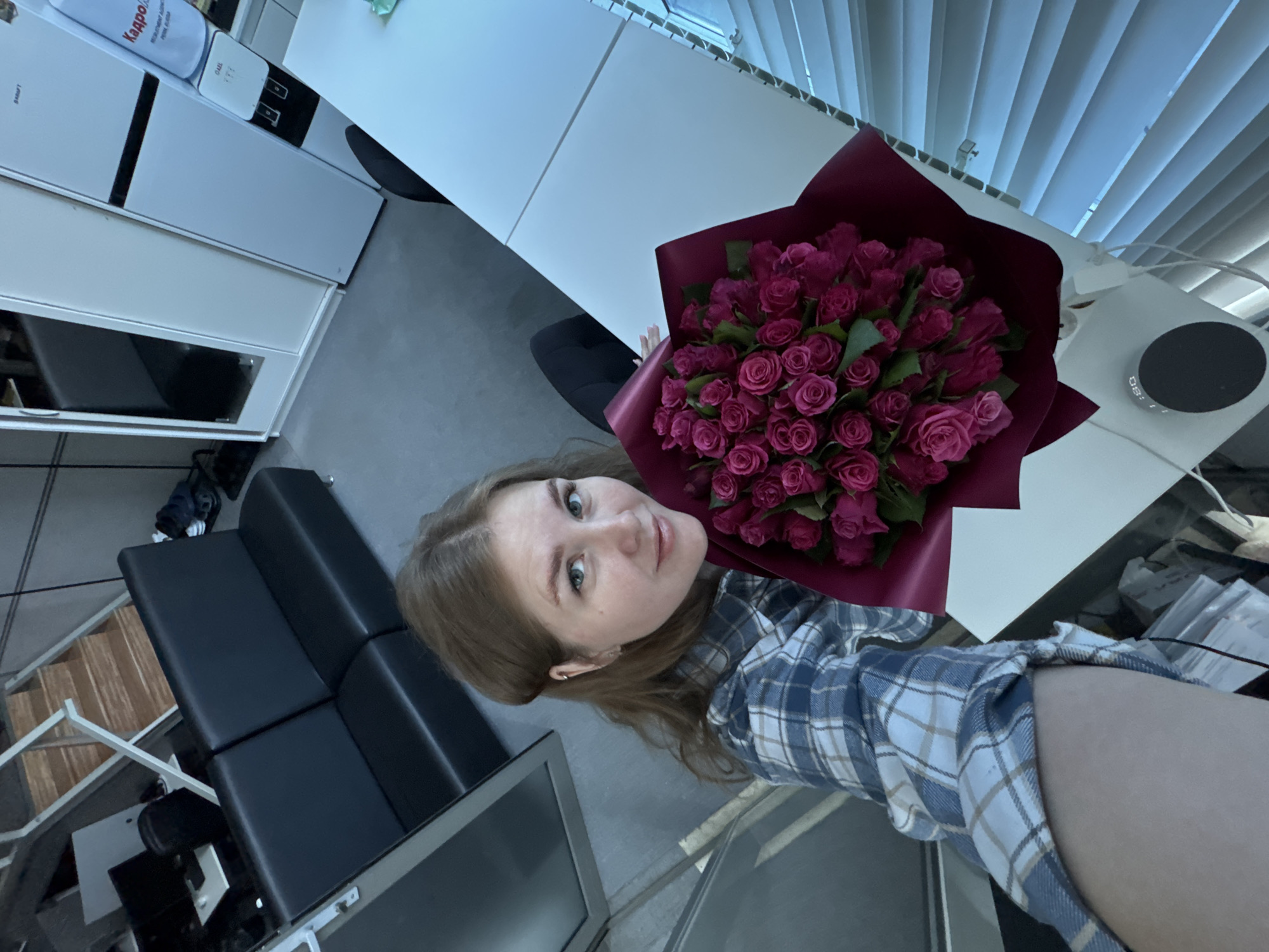 A young woman with shoulder-length brown hair and light blue eyes is taking a selfie while holding a large bouquet of pink roses. She is wearing a blue and white plaid shirt. The background appears to be an office or commercial space, with a black leather sofa and a staircase visible on the left. On the right, there is a white desk, a refrigerator, and a water cooler. The scene is lit with artificial light, and the overall impression is that of a casual moment captured in a professional setting. The image does not provide any specific clues to determine the exact location within the city of Perm, Russia.