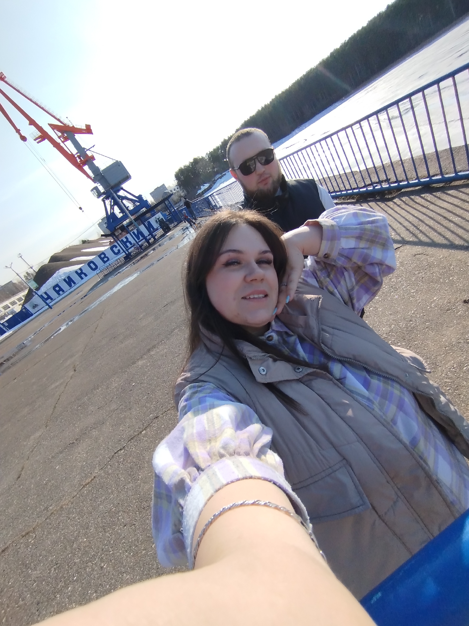A selfie shows a woman and a man standing on a paved area near the Chaikovsky port in Russia. Cranes and the name of the city (ЧАЙКОВСКИЙ) are visible in the background. The woman is in the foreground, wearing a plaid shirt and a vest. The man is behind her, wearing sunglasses and a vest. A body of water with ice and a tree line are also visible in the background, beyond a blue railing.