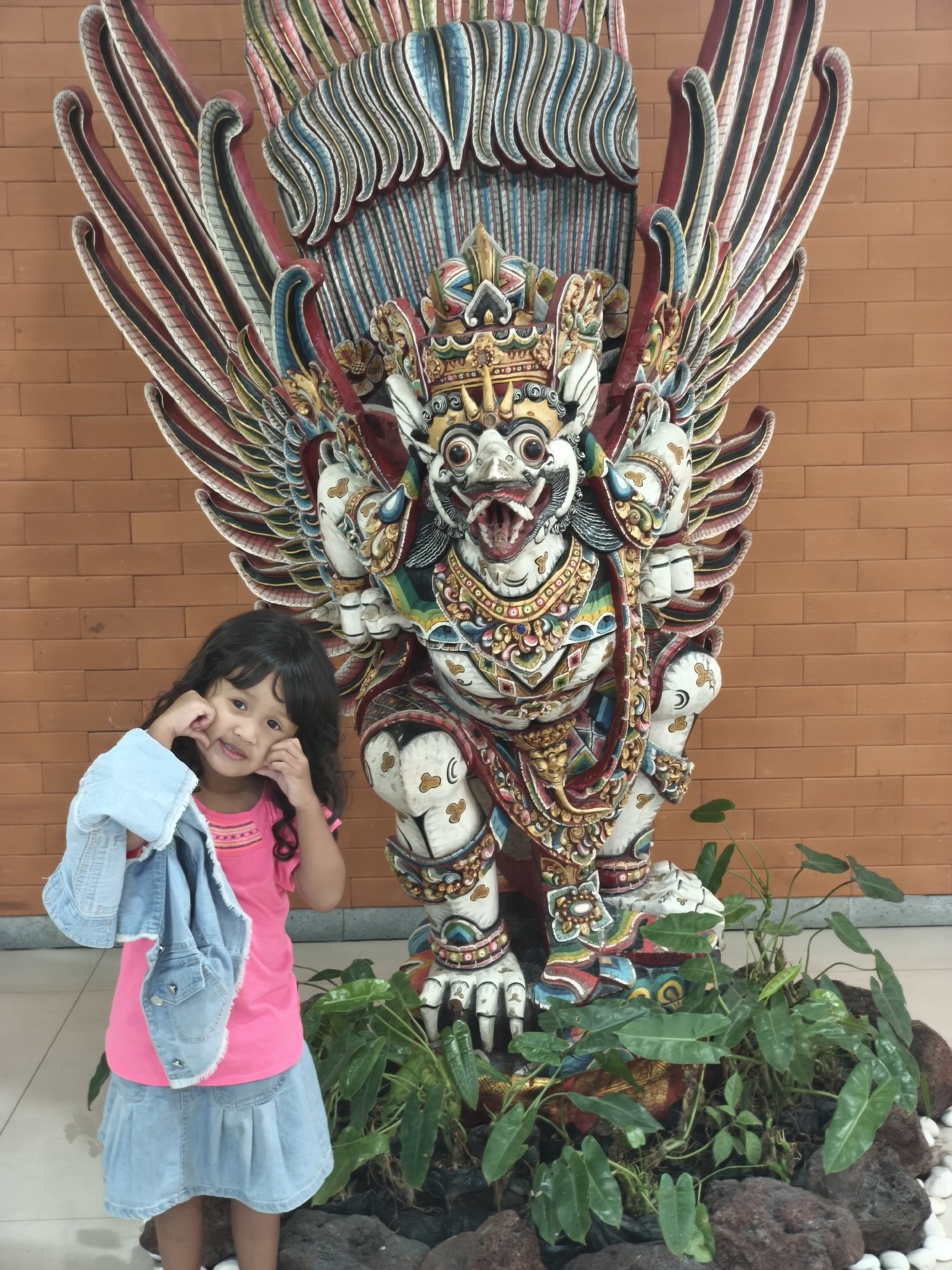 A young girl with dark, curly hair stands in front of a large, colorful statue of a mythical creature with wings. The statue is intricately carved and painted with vibrant hues of red, blue, yellow, and white. It depicts a fierce-looking being with a bird-like head, a crown, and multiple arms. The creature stands on a base surrounded by lush green plants and dark volcanic rocks.
The girl, wearing a bright pink t-shirt and a light blue denim skirt, holds a denim jacket over her arm. She is making a playful gesture with her hands near her cheeks, smiling at the camera. Her expression suggests a sense of joy and amusement. The background is a warm, textured brick wall. The lighting appears to be natural and even, suggesting it could be daytime, possibly indoors given the brick wall and tiled floor visible at the bottom of the frame. The overall scene is lively and captures a moment of childhood curiosity and engagement with cultural art.