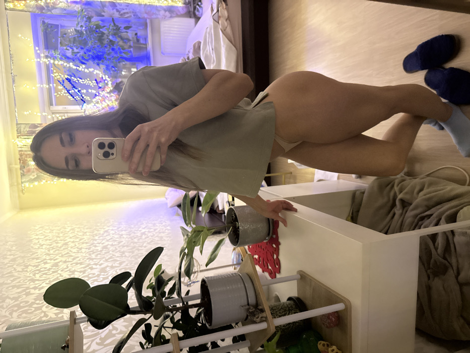 An indoor selfie captures a young woman in a bedroom or living space in Saint Petersburg, Russia, as reflected in a mirror. The woman, with long dark hair, is seen from the chest down, holding a light-colored smartphone with a prominent camera bump in her right hand to take the photo. She wears a loose-fitting, light grey or olive-colored short-sleeved top and light-colored underwear, revealing her bare legs and buttocks. Her left foot has a light blue sock, while her right foot is bare. Two dark blue fluffy slippers rest on the light-colored wooden floor behind her.
She strikes a dynamic pose, leaning her left hand on a white piece of furniture. Her right knee is bent, and her left leg is extended. The background features warm yellow artificial string lights decorating a window, with blue lights visible outside through the window, suggesting evening or nighttime. The walls are covered in patterned wallpaper. Several potted plants are arranged on a wooden stand with white railings and on other furniture. A white dresser or cabinet is visible, containing blankets or clothing. The overall ambiance is casual and personal.
