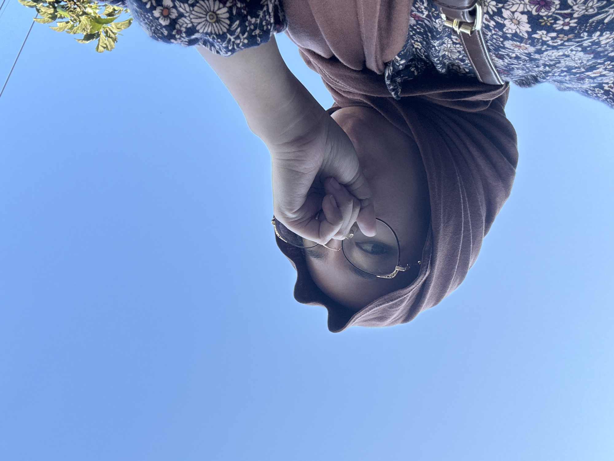 A woman wearing a brown hijab and glasses is shown from a low angle against a bright blue sky. Her right hand is raised, holding her glasses, with her fingers partially obscuring her face. She is also wearing a patterned dress and a brown belt. The image appears to have been taken outdoors, possibly on a sunny day, with the sky being the dominant feature of the background. There is a hint of green foliage in the upper right corner of the image. No specific location within Gadingrejo, Indonesia is discernible from the image itself.