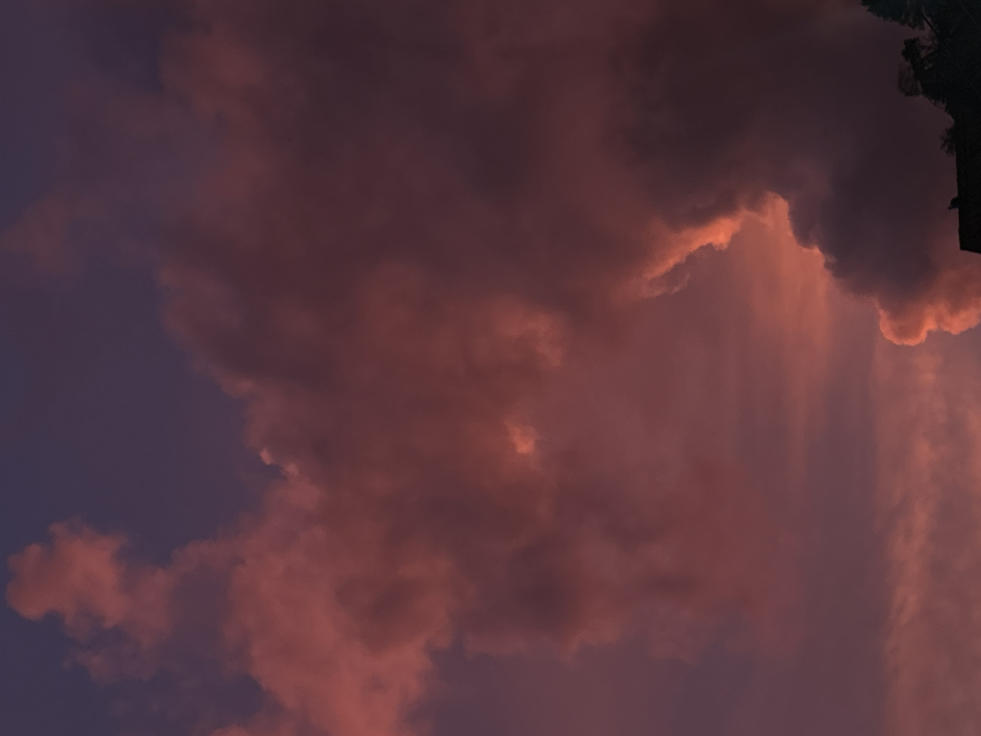 The image depicts a dramatic sky filled with billowing clouds tinged with hues of pink, purple, and orange, suggesting a sunset or sunrise. The clouds are dense and textured, with some areas brightly illuminated by the fading light, creating a sense of depth and volume. In the lower right corner, the silhouette of a rooftop and trees is visible, indicating an outdoor setting, possibly overlooking a landscape. The overall atmosphere is serene yet powerful, with the vibrant colors of the sky dominating the composition. There are no people or specific objects clearly identifiable other than the natural elements and the hint of human structures. The location is identified as Kathu, Thailand, implying this is a view of the sky above this region.