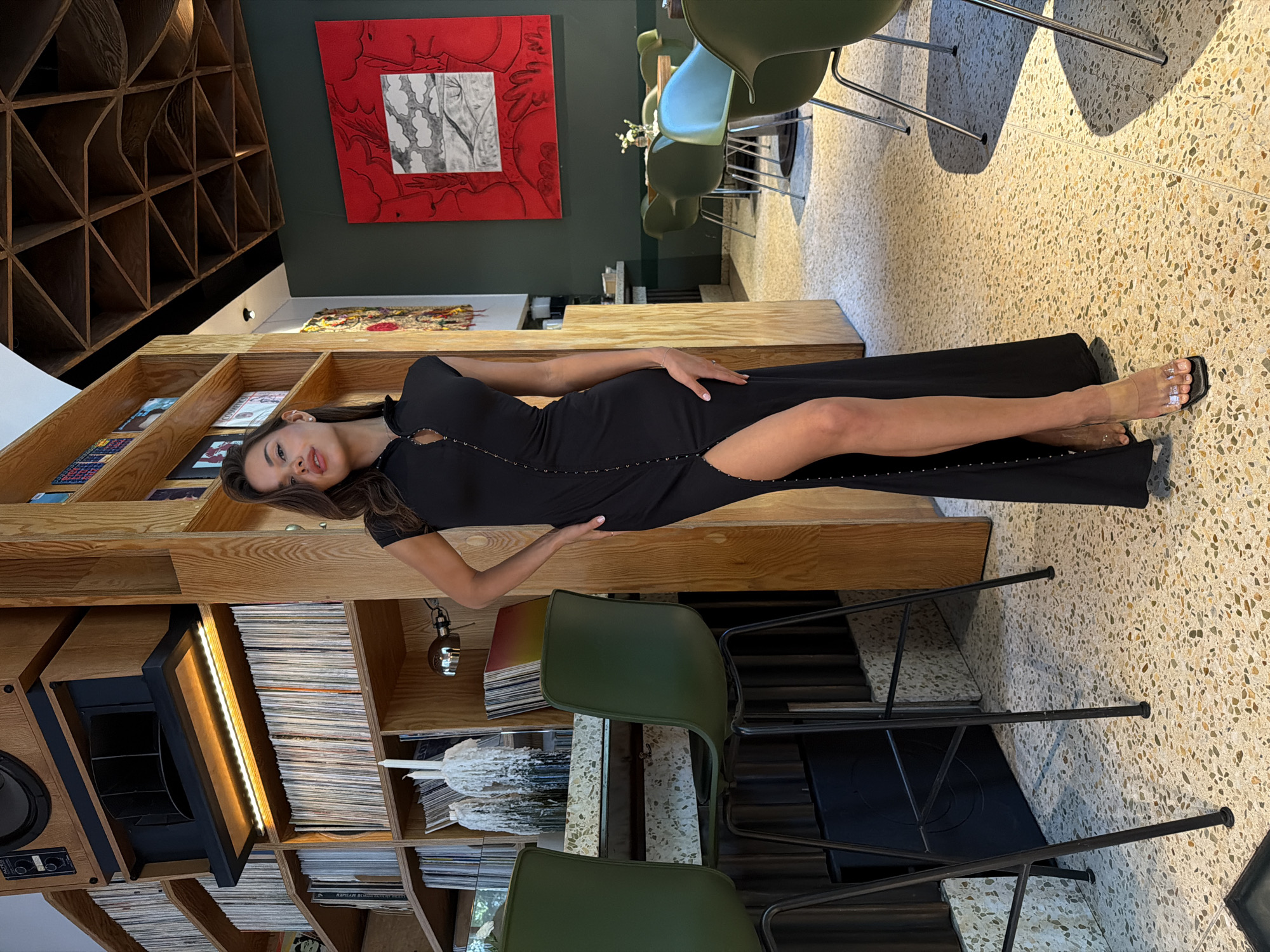 This image captures a young adult female standing within an interior space located in Moscow, Russia. The woman, positioned centrally and facing slightly left with a neutral expression, has long dark brown hair and is wearing a black, sleeveless maxi dress. The dress features a high collar and a central row of small, metallic hook-and-eye closures extending down the front to a high slit on the left side. She wears clear, open-toed high-heeled sandals. Her left hand rests on her hip.
The setting is characterized by its distinct decor. To the left, a tall wooden shelving unit displays numerous vinyl records. At the top of this unit, a large, vintage-style wooden audio speaker system with a horn-loaded component is mounted. Below the speaker, a linear LED light strip illuminates a shelf of records. Adjacent to this, a bar area features a light-colored, speckled terrazzo countertop. Two dark green bar stools with black metal frames are positioned at the counter. On the bar, two tall, white, heavily dripped candles stand next to a stack of records. A small chrome-finished wall lamp is affixed to a wooden pillar nearby.
The background to the right reveals a dining area with dark green walls. A large artwork, framed in red, hangs prominently on the wall, displaying a landscape with gray and white tones against a red-outlined backdrop. The ceiling above this area features a complex geometric pattern of dark wooden triangular panels. Multiple dark green chairs and one light blue chair, all with metal legs, are arranged around small, round tables. The entire floor of the visible area is covered in light-colored terrazzo with various embedded aggregate chips.