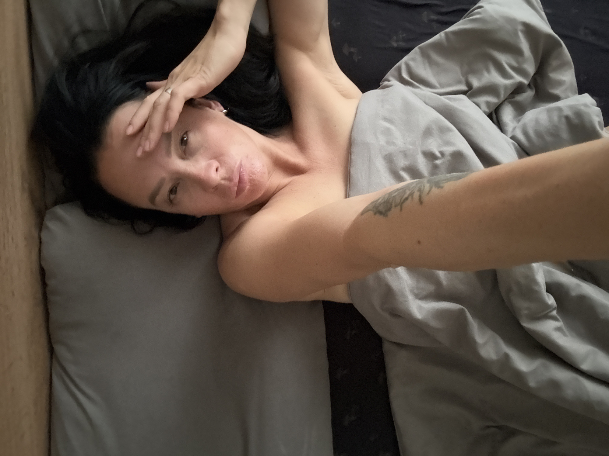 A woman with dark hair is lying in bed. She is under a gray blanket, with her arm extended above her head. Her face is partially obscured by her hand, but her eyes are open and she is looking towards the camera. The room is dimly lit, and the bed appears to be in a bedroom. The image is taken from a high angle, looking down at the woman. The location is not specified.