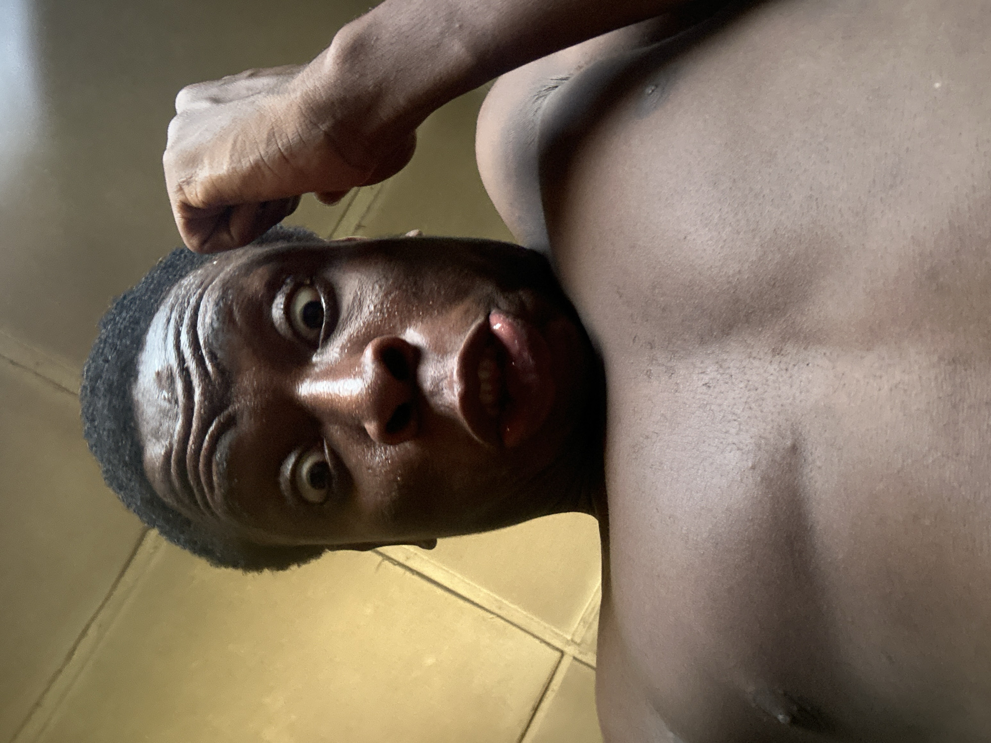 The image, rotated 90 degrees clockwise, features a close-up, low-angle shot of a person with dark skin. The individual, appearing to be male, is shirtless, revealing their upper chest, shoulder, and arm. Their head is tilted to the left, with short, dark, curly hair visible.
The person is looking directly into the camera with wide, intense eyes. Their mouth is slightly agape, revealing some teeth, which could suggest surprise, shock, or a strong emotional expression. Forehead creases are visible, adding to the intensity of the expression. The skin appears shiny, possibly from sweat or oil, highlighting the contours of the face and body.
One arm is raised, with the hand clenched into a fist, positioned above the person's head, near the top of the frame. The background is largely out of focus, consisting of indistinct, light-colored surfaces. In the upper right corner, a plain wall or ceiling is suggested, while a blurry, possibly tiled floor can be glimpsed below the person's head, indicating an indoor setting. The lighting is bright, creating distinct highlights on the skin. There is no visible text in the image.