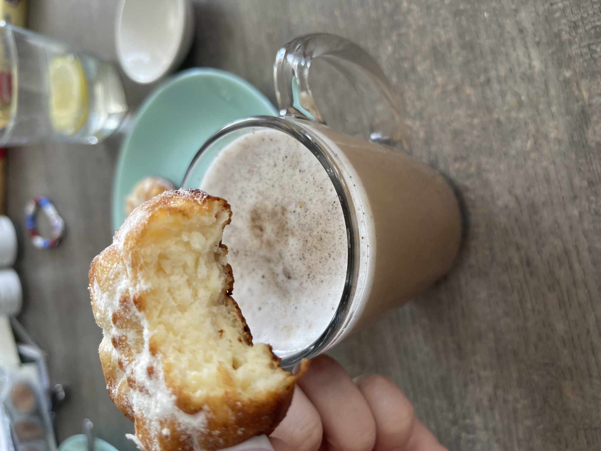 The image displays a close-up view of a person's hand holding a powdered pastry and a mug of coffee, set against a wooden surface. The pastry appears to be a fried dough confection, possibly a donut or churro, generously coated in powdered sugar and partially bitten into, revealing a soft, doughy interior. The coffee in the clear glass mug is topped with a frothy layer and dusted with cinnamon or cocoa powder.
In the blurred background, a pale green plate is visible, with what appears to be another pastry on it. To the left, a glass containing a drink with lemon slices and ice is also out of focus. Other objects like a white cup and some indistinct items are present but not clearly discernible.
The scene evokes a feeling of casual indulgence, perhaps a morning coffee break or a relaxed snack time. The lighting suggests it could be daytime. There is no visible text in the image. The overall impression is that of a simple, comforting moment. The location context is Blagoveshchensk, Russia.