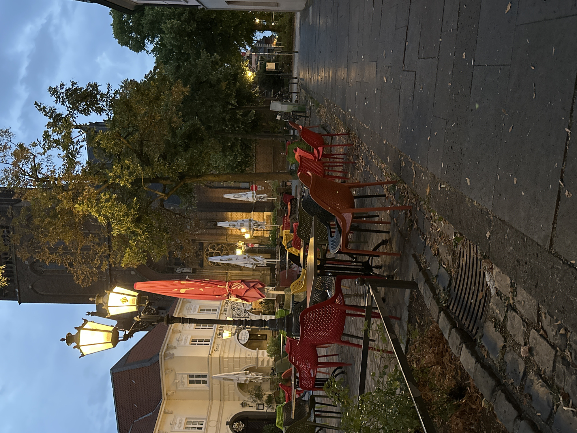 The image shows a deserted outdoor seating area of a cafe in Krefeld, Germany, in the evening. There are numerous tables and chairs arranged on a cobblestone patio. The chairs are brightly colored, in shades of red, orange, yellow, and black. A lamppost with two glowing lights stands to the left of the frame, illuminating the scene. In the background, a large building, possibly a church with a tall spire, is visible. Trees with autumnal foliage are scattered throughout the area. The sky is overcast and a pale blue. The overall impression is one of quietness and stillness, as if the cafe has recently closed or is about to open.