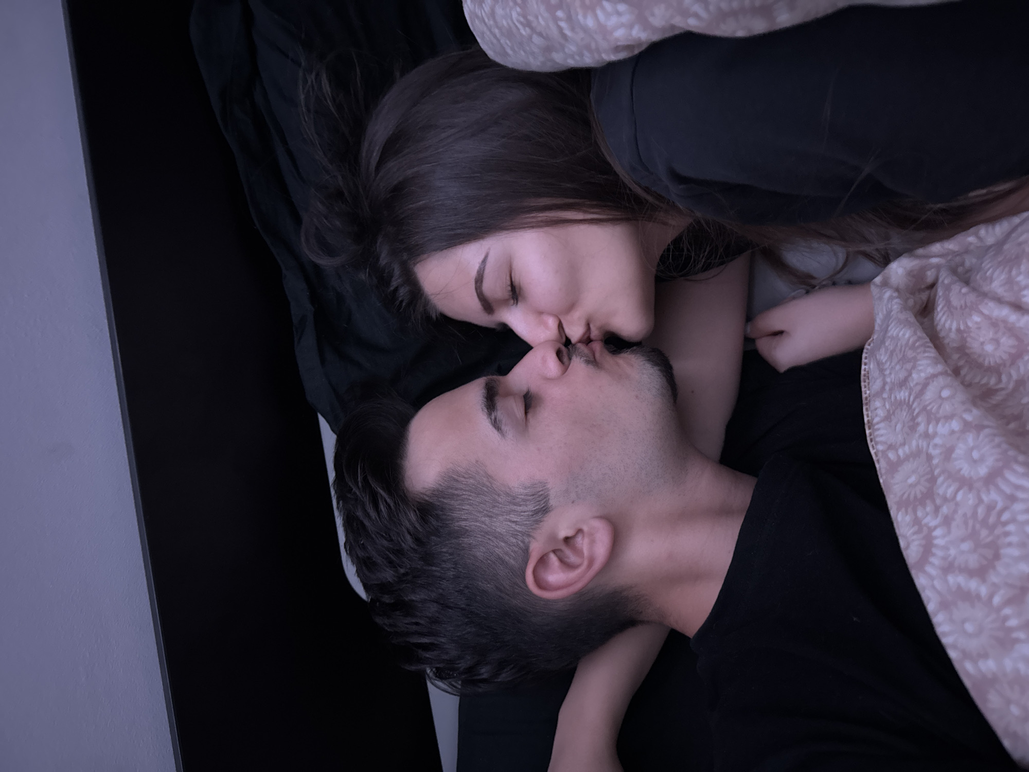 An overhead photograph captured in what is understood to be Sofia, Bulgaria, depicts two individuals, a male and a female, kissing in a bed. The scene is illuminated by dim, ambient light.
The male, positioned on the left side of the frame, lies on his back with his head turned to his right. He appears to be a young adult with short, dark hair, a light stubble on his chin and upper lip, and is wearing a dark (likely black) t-shirt. His left arm is bent, with his hand possibly positioned behind his head or under a pillow. His eyes are closed.
The female, positioned on the right side of the frame, lies on her left side, facing the male. She also appears to be a young adult with long, dark hair. She is wearing a dark, long-sleeved garment, possibly a t-shirt or hoodie. Her eyes are closed.
The two individuals' lips are pressed together in a kiss.
They are situated in a bed, covered partially by a light-colored blanket or duvet featuring a repetitive patterned design of circular or floral motifs. Dark bedding, possibly sheets or a fitted sheet, is visible beneath them and extending towards the top of the bed frame. A dark, possibly black, headboard or bed frame is visible behind their heads, with a light purple-grey wall above it. The overall impression is an intimate, private setting, likely a bedroom.