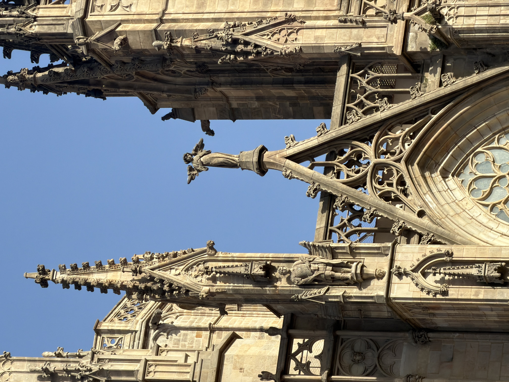 The image shows an architectural detail of a Gothic cathedral, likely in Barcelona, Spain, given the ornate stone carvings and sculptures characteristic of Catalan Gothic architecture. The scene is dominated by intricately carved stone elements, including spires, pinnacles, and decorative tracery. Prominently featured are two sculptures: a winged angel standing atop a spire on the left, and a male figure in armor, resembling a warrior or saint, embedded in the facade on the left side of the image. Gargoyles are visible on the upper sections of the cathedral's architecture. The stonework is weathered and appears to be made of sandstone or a similar material, bathed in warm sunlight against a clear blue sky. The overall impression is one of historical grandeur and detailed craftsmanship.