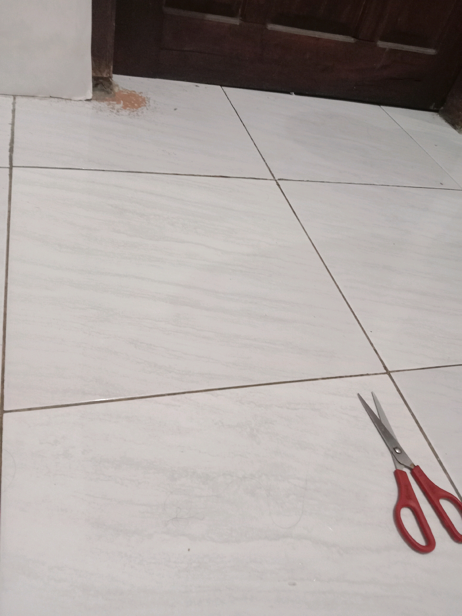 A pair of red-handled scissors lies open on a light-colored tiled floor, near the bottom right corner of the image. The tiles are laid in a grid pattern with dark grout lines. In the upper left corner, a small area of the floor shows a patch of brownish-orange discoloration next to a vertical wooden surface. The background consists of a dark wooden structure, possibly part of a door or cabinet, in the upper portion of the image. The scene appears to be indoors, in a room with tiled flooring. The location in Brangsong, Indonesia, is not visually determinable from the image.