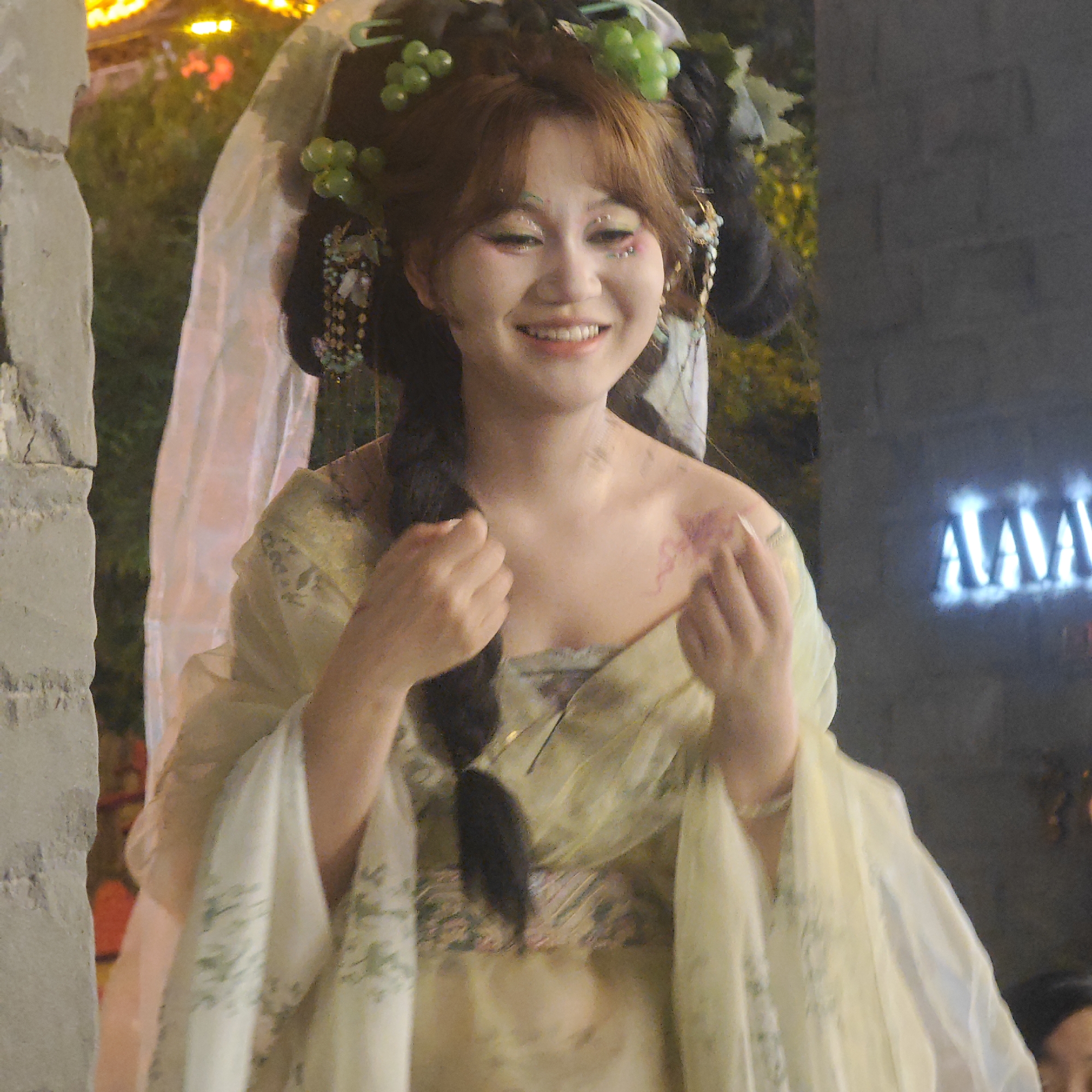 A smiling young woman in traditional dress and elaborate makeup stands in an outdoor setting in Yuzhong District, China. She wears a light yellow and green dress with floral patterns and a draped overlay. Her hair is styled in an intricate updo, adorned with green grapes and other decorations. A long black braid hangs down her shoulder. The woman's face is painted with colorful eye makeup and sparkles. She is framed by a stone structure on the left and a dark wall with neon lettering on the right. Behind her, blurred lights and greenery suggest a public area. She holds her hands near her chest.