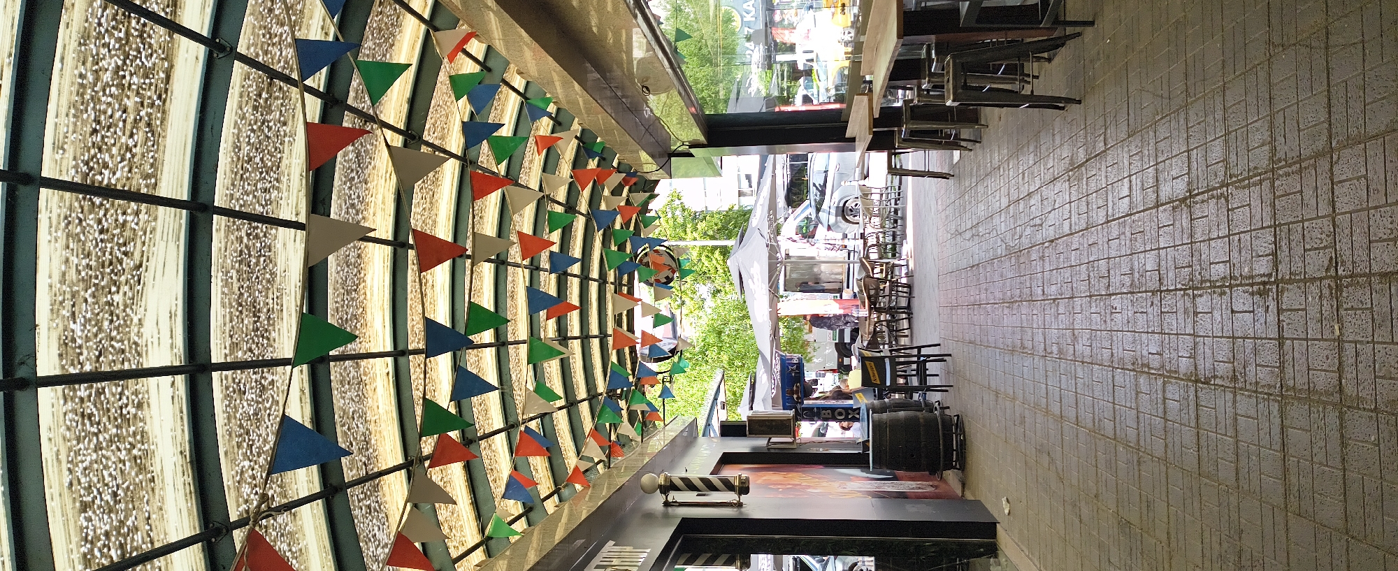 This outdoor scene is set in Sofia, Bulgaria, on a pedestrian walkway constructed of rectangular, grey paving stones with visible dirt accumulation in the grooves. The image is oriented so that the walkway runs roughly from the upper right to the lower right corner.
On the left side is the exterior of a building. A series of colored triangular pennants (blue, green, orange, and beige) are strung along a metal grid structure that covers the storefront's overhang. Beneath this overhang, a barber pole indicates a barbershop. A black trash can and several black barrels are situated near the store.
The walkway to the right is lined with outdoor seating for a café or restaurant. Wooden tables are placed along the walkway, each with multiple chairs. Further down the walkway, more tables are visible, with some covered by a white canopy. People are present, interacting near the tables beneath the canopy.
Reflections in the building's windows indicate a street scene with cars and greenery beyond.