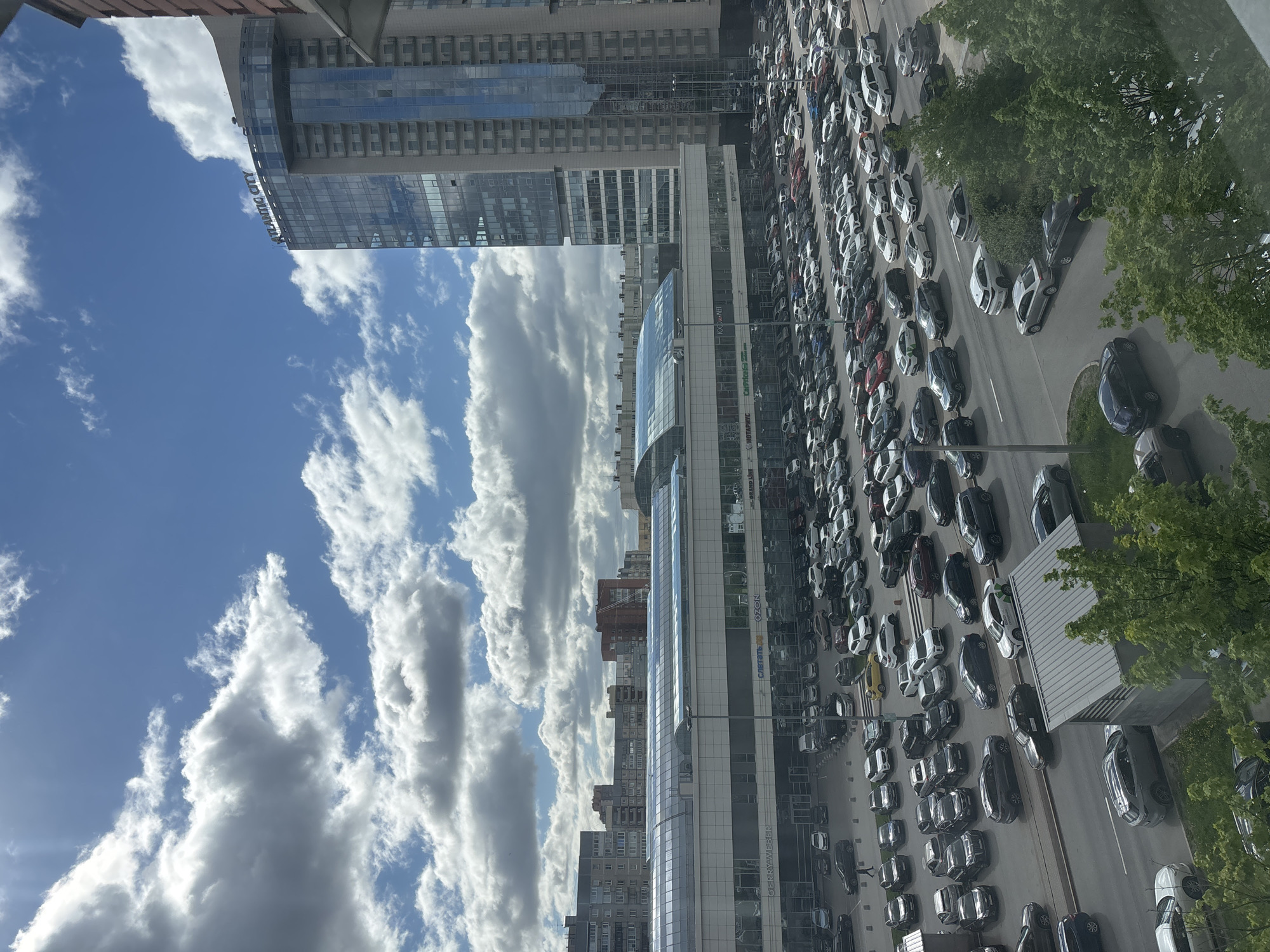 The image shows a congested street in Saint Petersburg, Russia. A dense line of cars fills the road, viewed from an elevated position, possibly a building. Modern buildings with glass facades line the street, reflecting the sky. The sky is filled with cumulus clouds. Trees are present along the edge of the road. There are no people clearly visible.