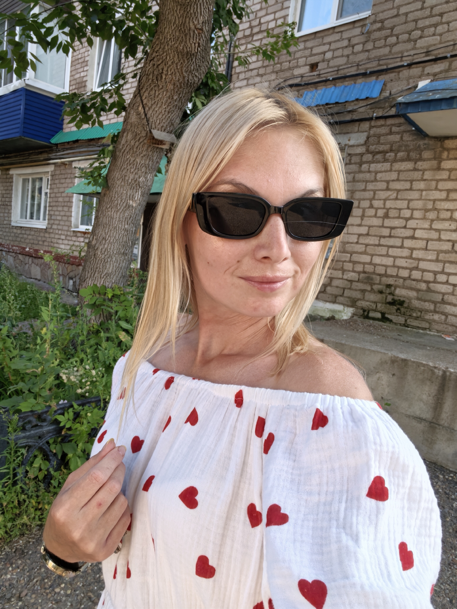 A blonde woman wearing sunglasses and a white off-the-shoulder shirt with red hearts is standing outdoors in front of a brick building and a tree. She is looking towards the camera with a slight smile. The background includes greenery, a blue balcony, and windows. The overall scene suggests a casual, daytime setting in a residential area, likely in Sterlitamak, Russia, given the architectural style.