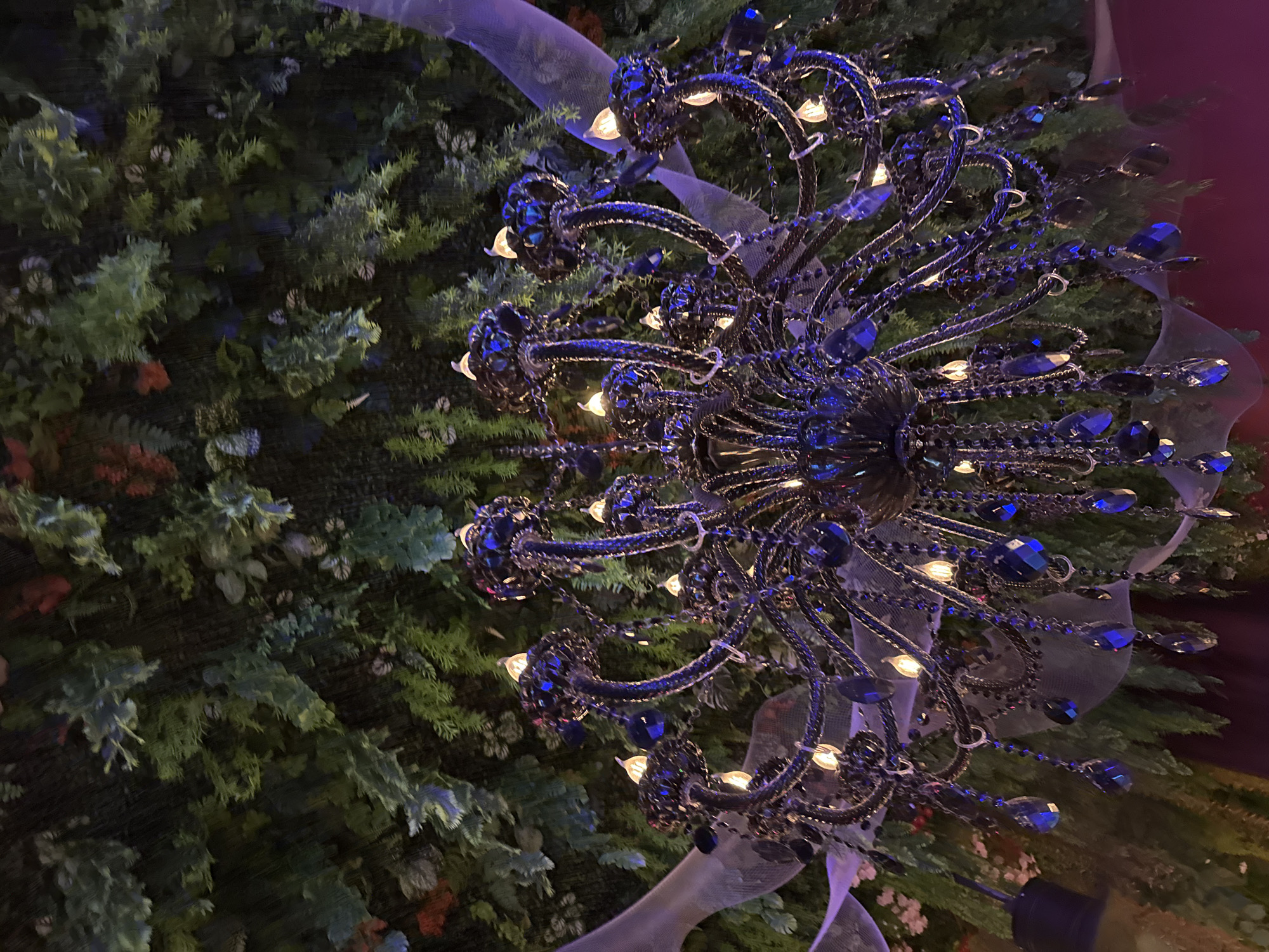 Captured from below, the image features a dark, multi-tiered chandelier adorned with blue crystals and small lights, set against a backdrop of dense, green foliage. Sheer, light-colored ribbons are interwoven among the chandelier's arms and the foliage. The scene suggests an indoor setting, possibly a restaurant or event space in Baan Chaweng, Thailand.