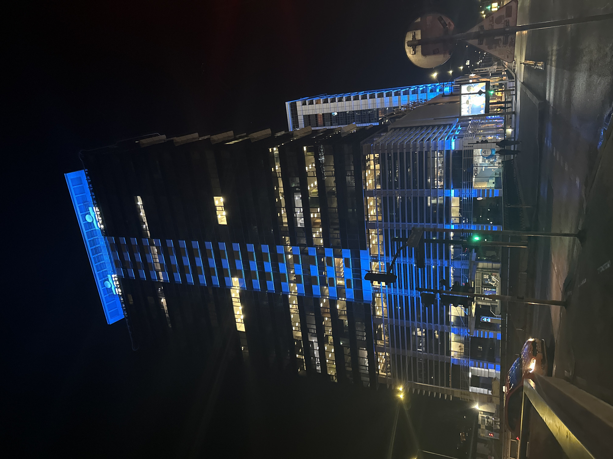 This night-time image captures a modern, tall building in an urban setting, likely the Vivacom Tower in Sofia, Bulgaria. The building dominates the left side of the frame, illuminated with striking blue lights on large sections of its exterior facade, creating a distinct grid pattern. Many of its interior windows also glow with a warm yellow light. Vertically displayed in bright blue light on the upper left side of the building is the text "VIVACOM," with what appears to be its logo below it.
On the right side of the frame, a wet street or sidewalk reflects the artificial light sources, suggesting recent rain. Several indistinct human figures are visible on the sidewalk near the base of the building. A large, circular convex traffic mirror is mounted on a pole towards the top right, and traffic lights are visible further down the street. A car, possibly a taxi given its roof light, is partially visible on the far right. Streetlights cast pools of light onto the damp ground, contributing to the vibrant atmosphere of the city at night.