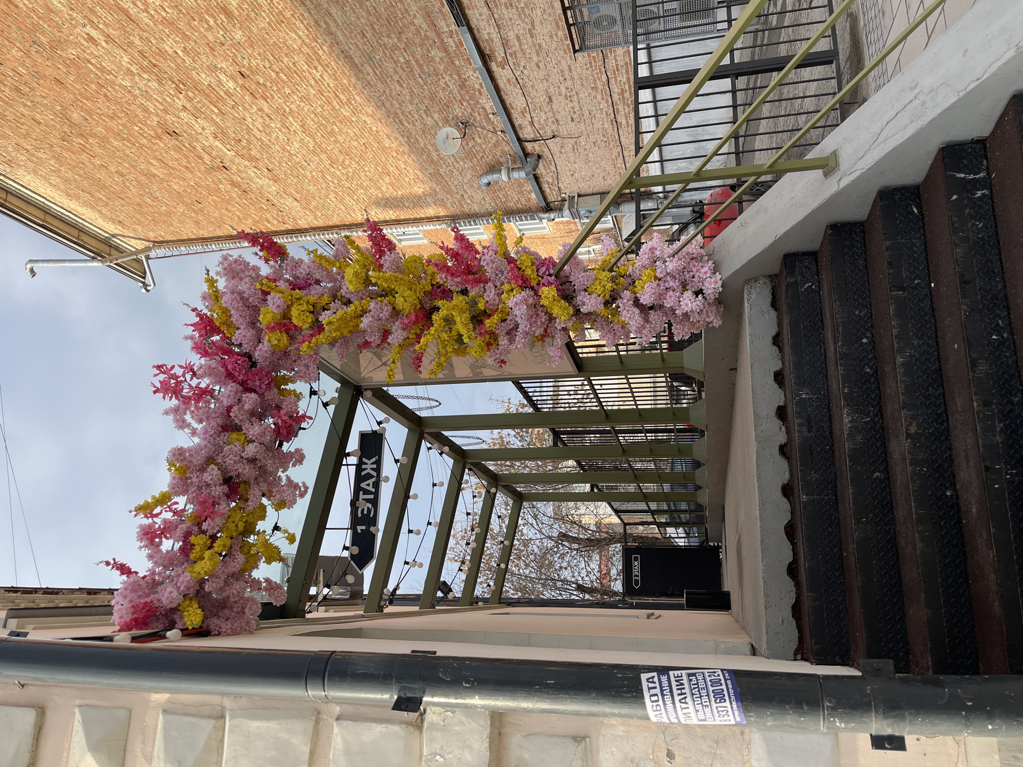 The image depicts a street scene, likely an entrance to a building. A floral archway composed of pink and yellow flowers frames the entryway. The building's facade is a mix of brick and painted concrete. A sign written in Russian, partially obscured, indicates "1 ЭТАЖ" and "2 ЭТАЖ", suggesting multiple floors within the building. A metal awning covers the entrance, and nearby stairs lead upwards. A pipe with a sticker bearing Russian text and a phone number is visible at the bottom. The location is suggested to be Izhevsk, Russia.