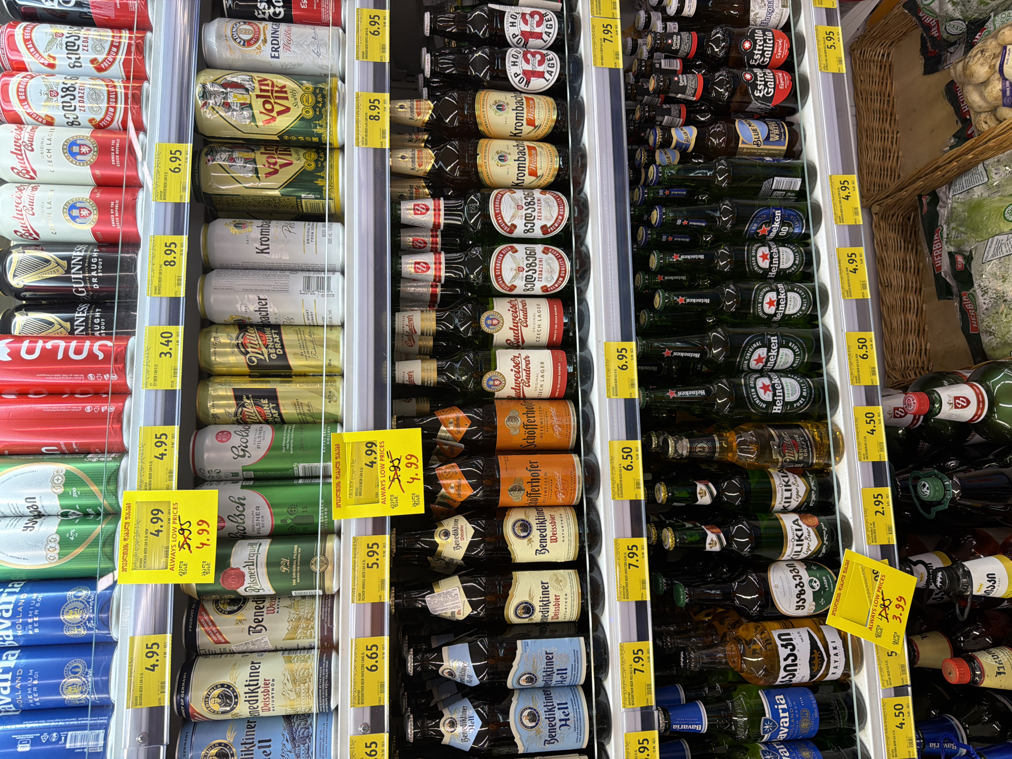 The image displays a retail shelf stocked with a variety of alcoholic beverages, primarily beer, in a supermarket environment. The arrangement suggests a commercial setting, possibly in Tbilisi, Georgia, given the presence of Georgian language text on some of the product labels and price tags.
The shelf is organized into several tiers, with canned beverages on the upper shelves and bottled beverages on the middle and lower shelves. Price tags are visible beneath many of the products, indicating individual item pricing. Some price tags are in Georgian Lari, and some are in euros, which is common in Georgia.
On the top shelf, various brands of beer cans are visible, including brands with labels that appear to be in Georgian. There are also cans of Guinness, Budweiser, and other international brands.
The middle shelf features bottled beers. Prominent brands include "Benediktiner Hell," "Benediktiner Weissbier," and "Schöfferhofer." These bottles are predominantly glass and vary in color from clear to amber.
The bottom shelf displays more bottled beverages, including brands like "Bavaria," "Kayaki," "Kilikia," and "Miller Genuine Draft." Some of these bottles are also green glass, typical for beer. The very bottom of the image shows the tops of more bottles, and some wicker baskets likely used for storage or display.
A yellow price tag with Georgian text and "ALWAYS LOW PRICES" written in English is prominently displayed in the middle section, highlighting a discounted price of 3.99. This indicates a promotional sale.
There are no people visible in the image, and the focus is solely on the product display. The lighting appears to be standard supermarket fluorescent lighting, creating a well-lit but somewhat sterile environment. The overall scene is a typical supermarket aisle dedicated to alcoholic beverages.