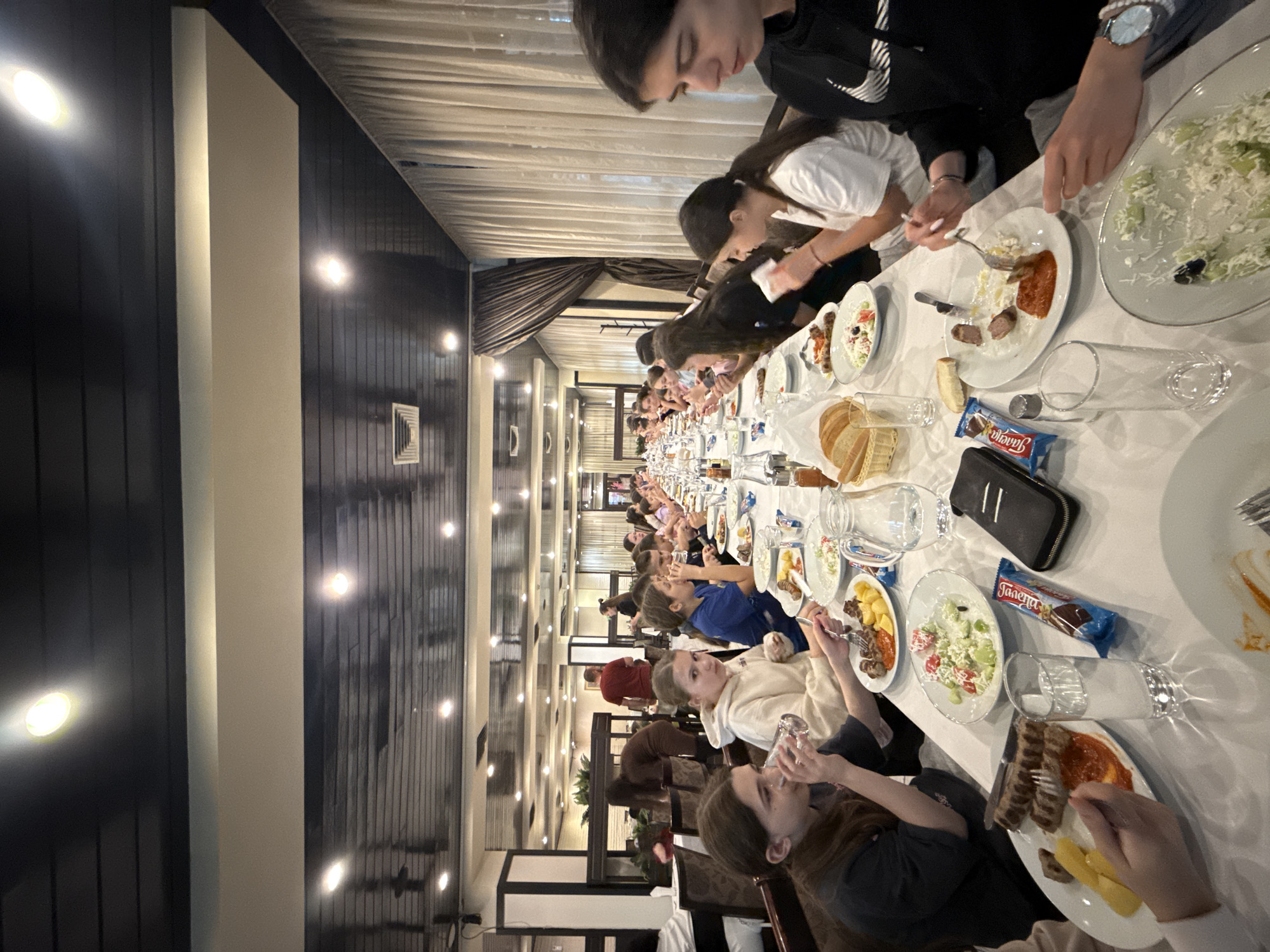 A long, extended table in a restaurant setting in Etropole, Bulgaria, is surrounded by a large group of people who are eating. The table is set with white tablecloths, plates of food, glasses, and bottles of water. Individual food items on the plates appear to be a mix of grilled meat, sliced vegetables, and salads. Some individuals are drinking from their glasses, while others are actively eating with utensils. Chocolate bar wrappers are visible scattered on the table. Overhead lighting fixtures illuminate the room, with a row of vertical light panels visible on the ceiling. The walls have a dark wood paneling at the top, with white sections below. The background features a curtained area and additional tables, indicating a large dining space.