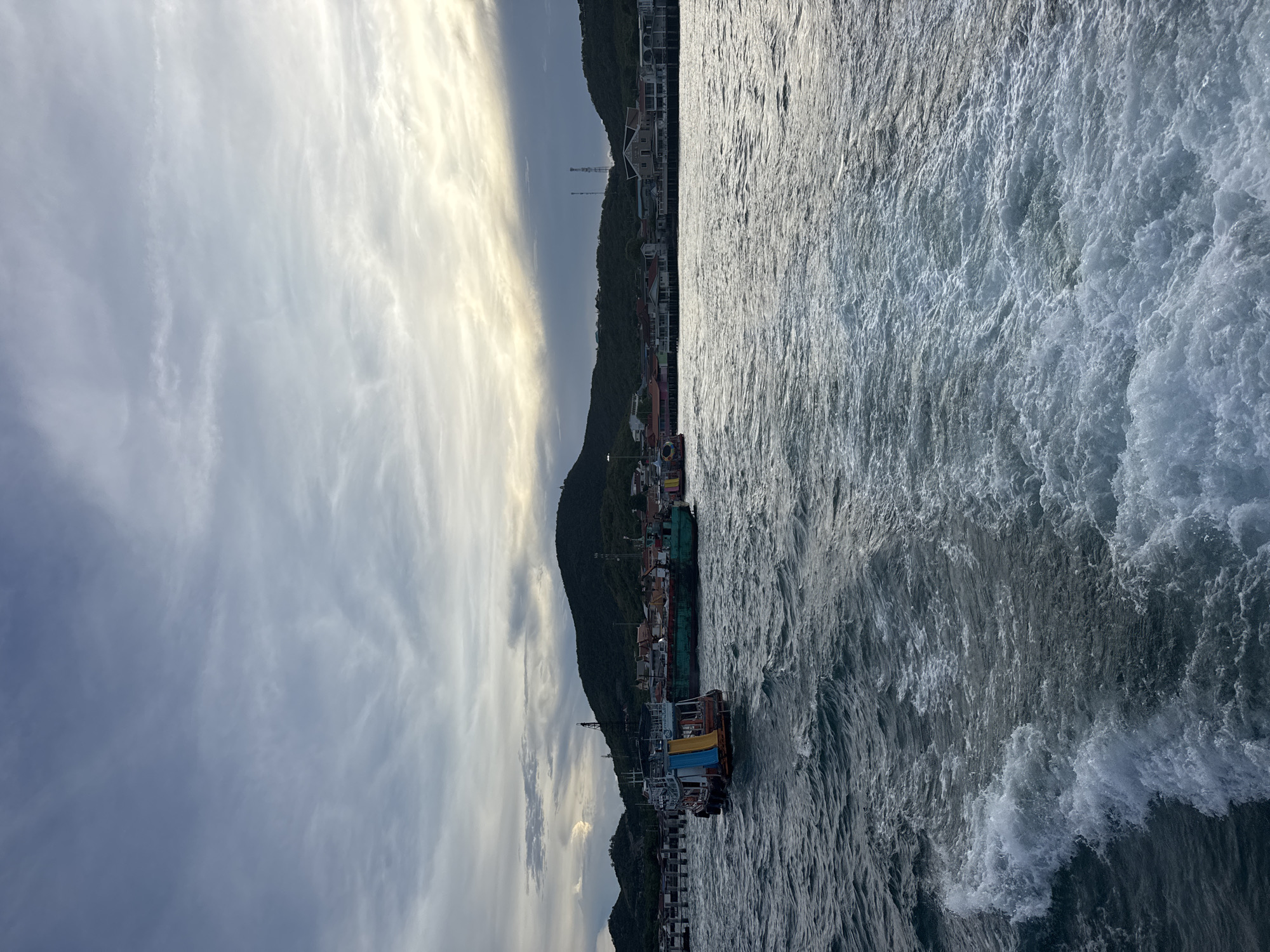 The image shows a waterfront scene in Pattaya City, Thailand. The composition is divided between sky, land, and water. A cloudy sky takes up the upper portion. A coastal hillside with buildings and infrastructure runs along the middle. In the foreground is turbulent water, suggesting movement. A pier with colorful boats is visible along the hillside.