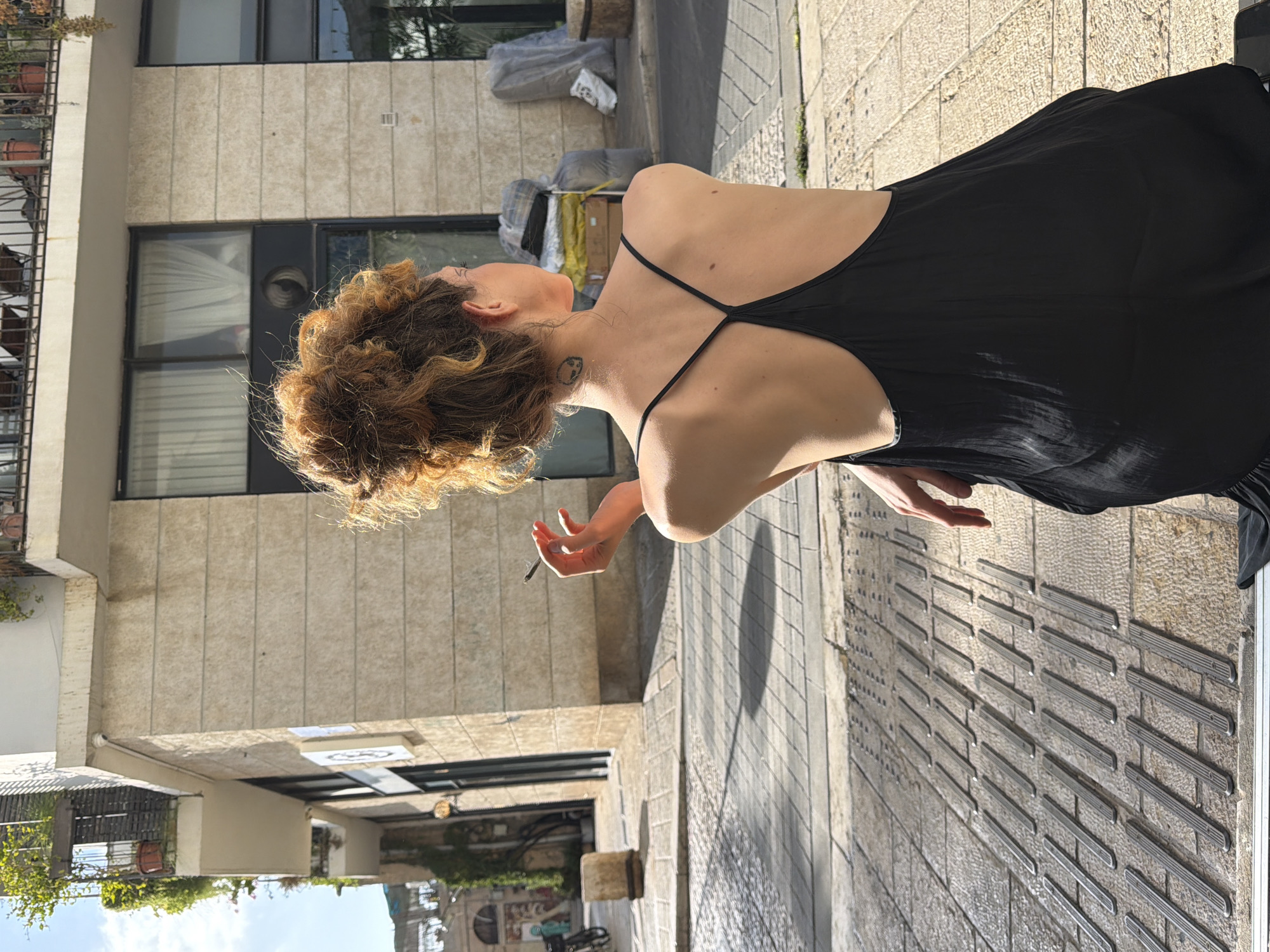 A woman with her back to the camera, wearing a black spaghetti strap dress, sits on a textured grey surface. Her auburn hair is styled in a messy bun. She holds a lit cigarette in her right hand, which is raised to her mouth. A small, dark tattoo of a skull is visible on the back of her neck. The background shows a building with a beige stone facade, balconies with plants, and windows. The ground is paved with patterned stone tiles. In the distance, there are more buildings, a bicycle, and some stacked items wrapped in plastic. The scene is outdoors in a city, likely Tel-Aviv, Israel, given the architecture and paving style often found there. The lighting suggests it is daytime, with sunlight casting shadows on the ground.