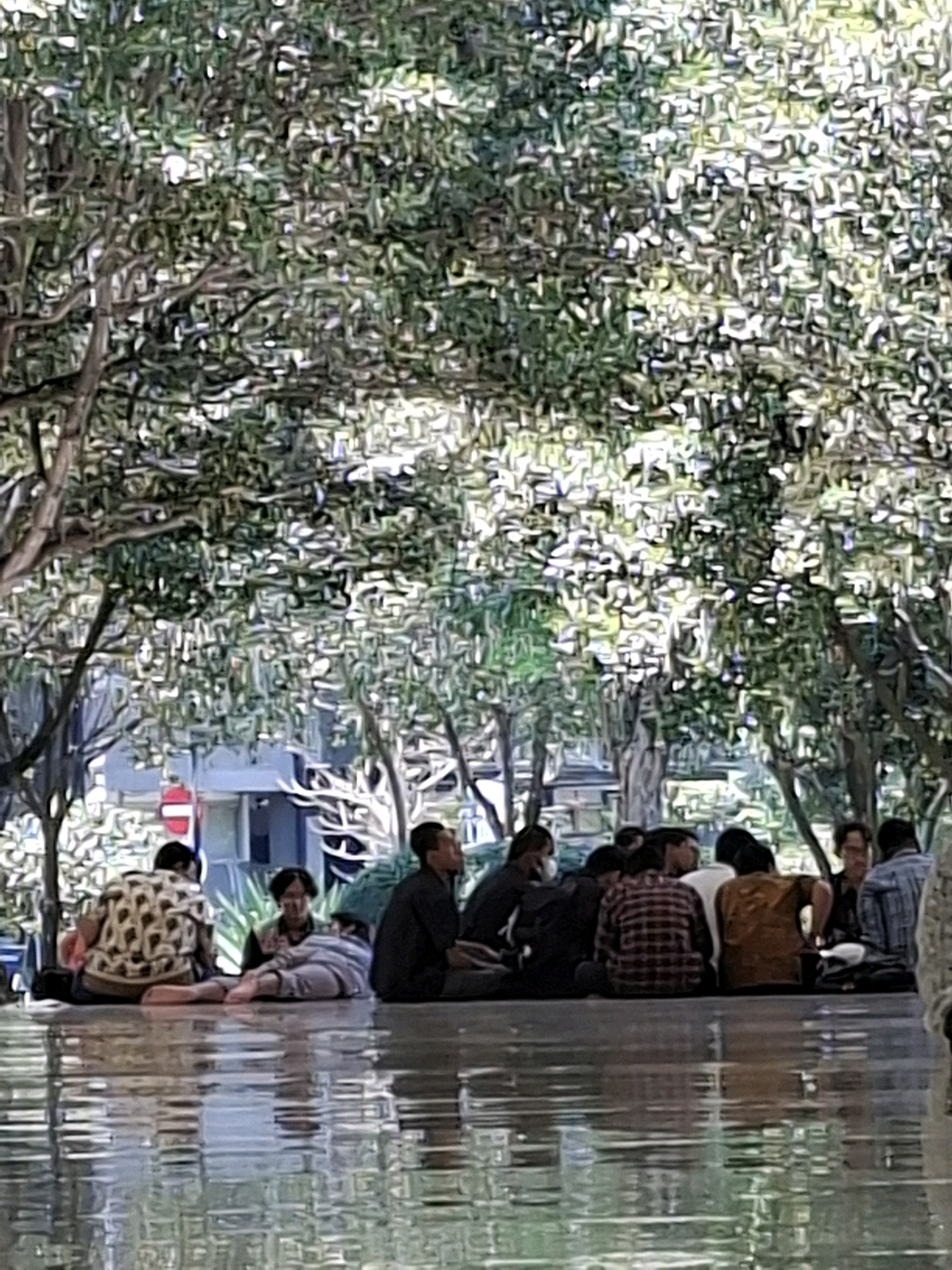 A group of people are sitting on the wet tiled floor beneath a large tree with dense foliage. The ground appears to be reflective, suggesting it may be wet from recent rain or a water feature. The people are casually dressed, and some are interacting with each other, while others are looking away. The background shows some buildings and trees, hinting at an outdoor urban environment. The scene is illuminated by natural light filtering through the tree canopy. The presence of a red and white sign with a red circle and line indicates a no-entry or prohibition symbol, suggesting a specific area within a public space. The overall impression is one of a relaxed gathering in a shaded outdoor area within Surabaya, Indonesia, possibly a park or a plaza.