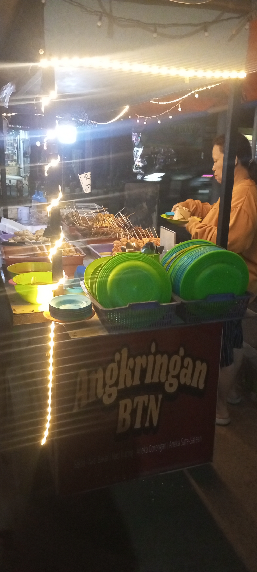 In Setu, Indonesia, at night, a food stall is depicted. A woman, wearing an orange long-sleeved shirt and striped pants, is shown from the side, preparing food for a customer. In front of her, several green plastic plates are stacked in baskets. To the left, a green bowl contains liquid, likely sauce, and there are other bowls and plates. Various skewers of food, including what appear to be fried snacks and meat on sticks, are displayed in containers behind the woman. A sign on the front of the stall reads "Angkringan BTN," with smaller text below indicating food options such as "Sedia Nasi Bakar, Nasi Kuhing, Aneka Gorengan, Aneka Sate-Satean." The stall is illuminated by strings of warm-toned lights. The overall environment suggests a bustling street food scene.