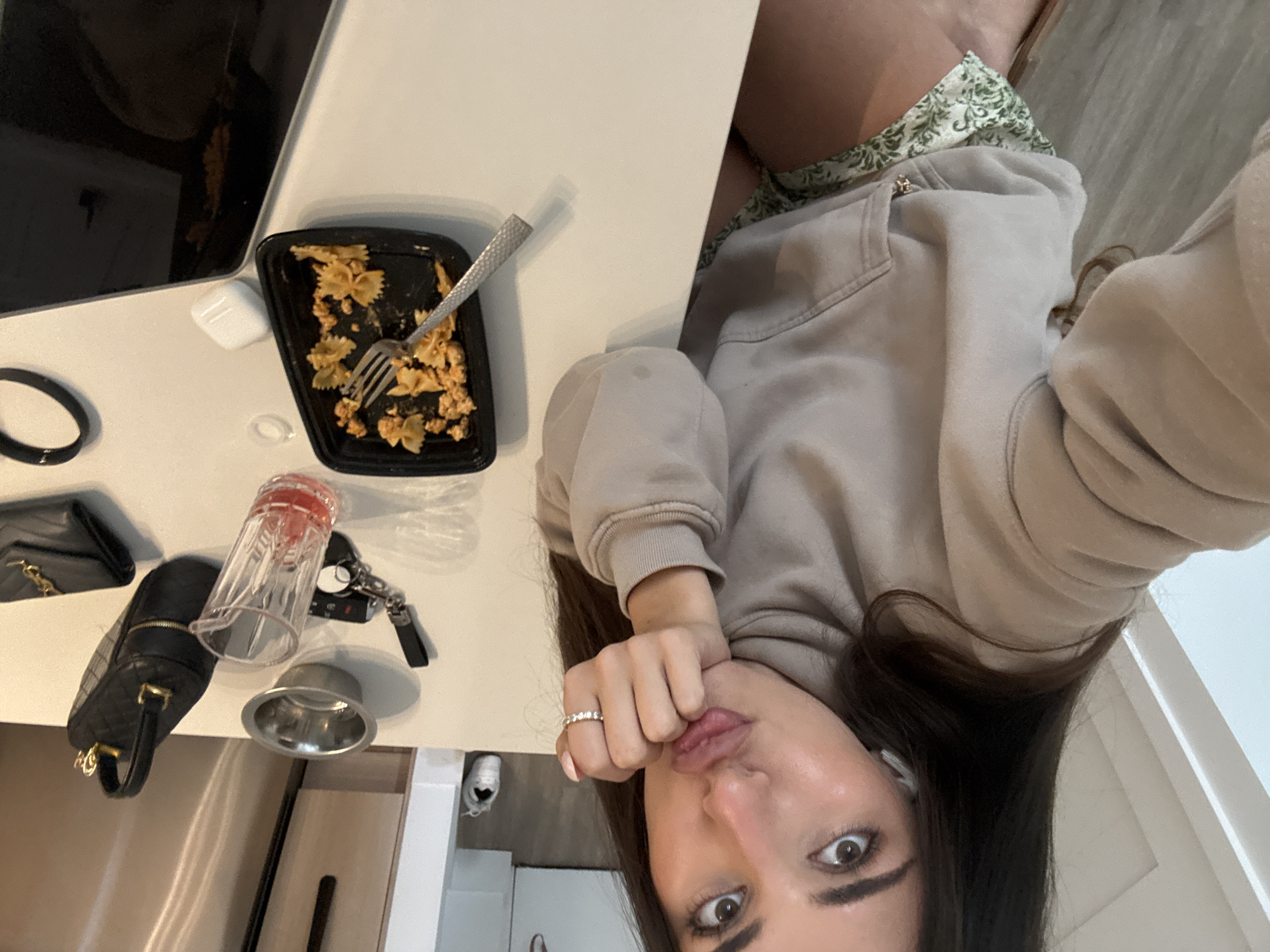 A woman with long dark hair is sitting at a table, holding her chin with her hand, and posing for a selfie. She is wearing a light-colored hoodie and patterned shorts. On the table in front of her is a black plastic container with pasta and a fork, a clear glass with pink liquid, keys, a black handbag, a black wallet, and a silver dog bowl. A pair of white earbuds are also on the table. The scene appears to be indoors, possibly in a kitchen or dining area.