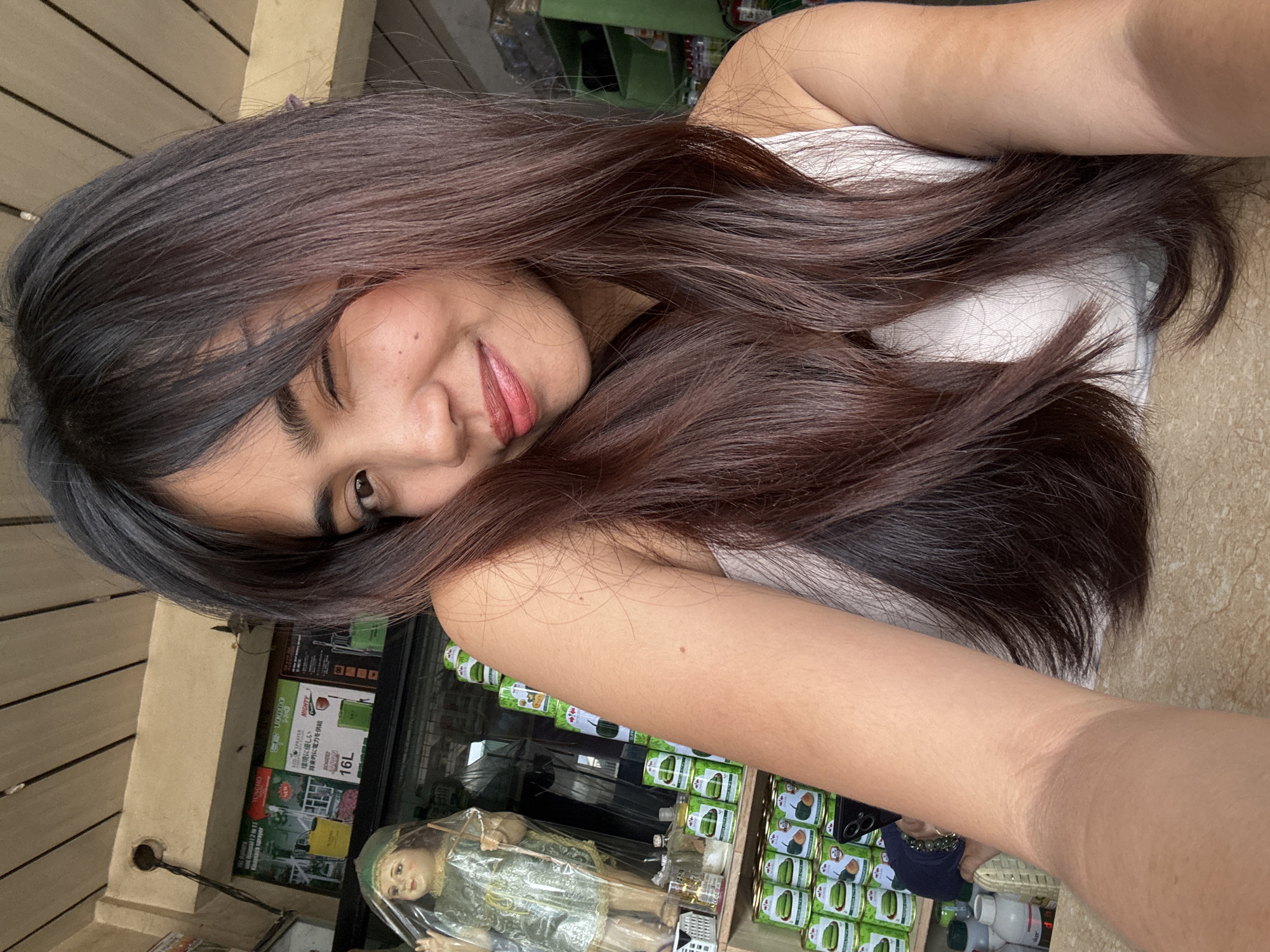 A young woman with long, dark brown hair and bangs winks at the camera, her head tilted and her right arm resting on her chest. She is wearing a white tank top. Behind her, shelves are filled with canned goods, plastic bottles, and a doll. The scene appears to be an indoor market or store, possibly in Tagum City, Philippines, given the presence of what seem to be agricultural products and local items. The lighting suggests it is daytime.