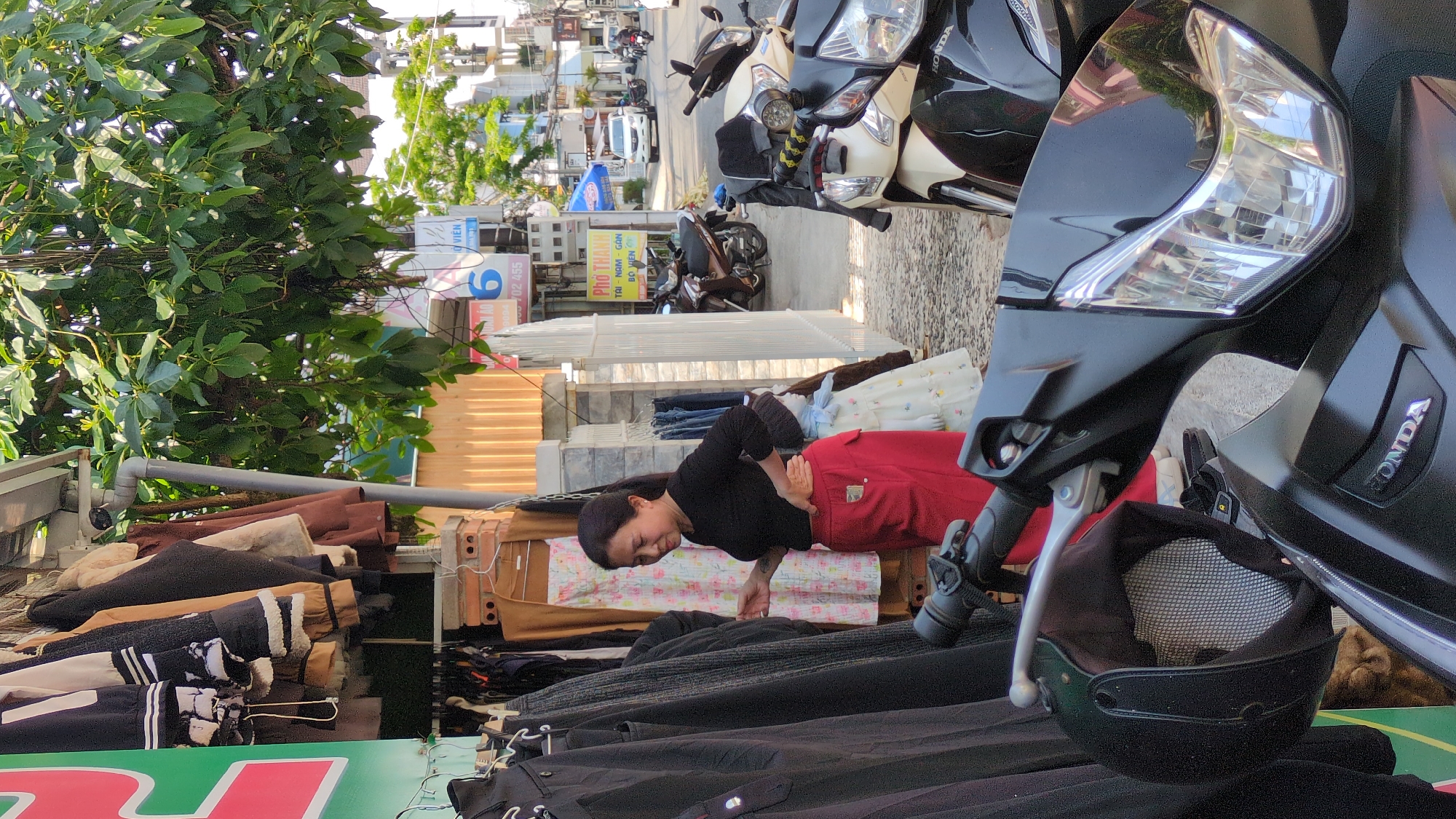 The image shows a street scene in what appears to be Da Lat, Vietnam. A woman wearing a black shirt and red pants is bent over, examining clothes hanging on a rack in front of a shop. The clothes on the rack include jackets and other garments. The street scene includes parked motorbikes, a common mode of transportation in Vietnam. There is a sign visible for "Pho Thanh Tai - Nam - Gan Bo Vien," suggesting the presence of a pho restaurant nearby. Green foliage is seen on the left side of the frame, possibly a tree or shrub lining the street. The perspective is from a low angle, with a motorbike in the foreground on the right bearing the Honda logo. Buildings and signs of a bustling street are visible in the background.