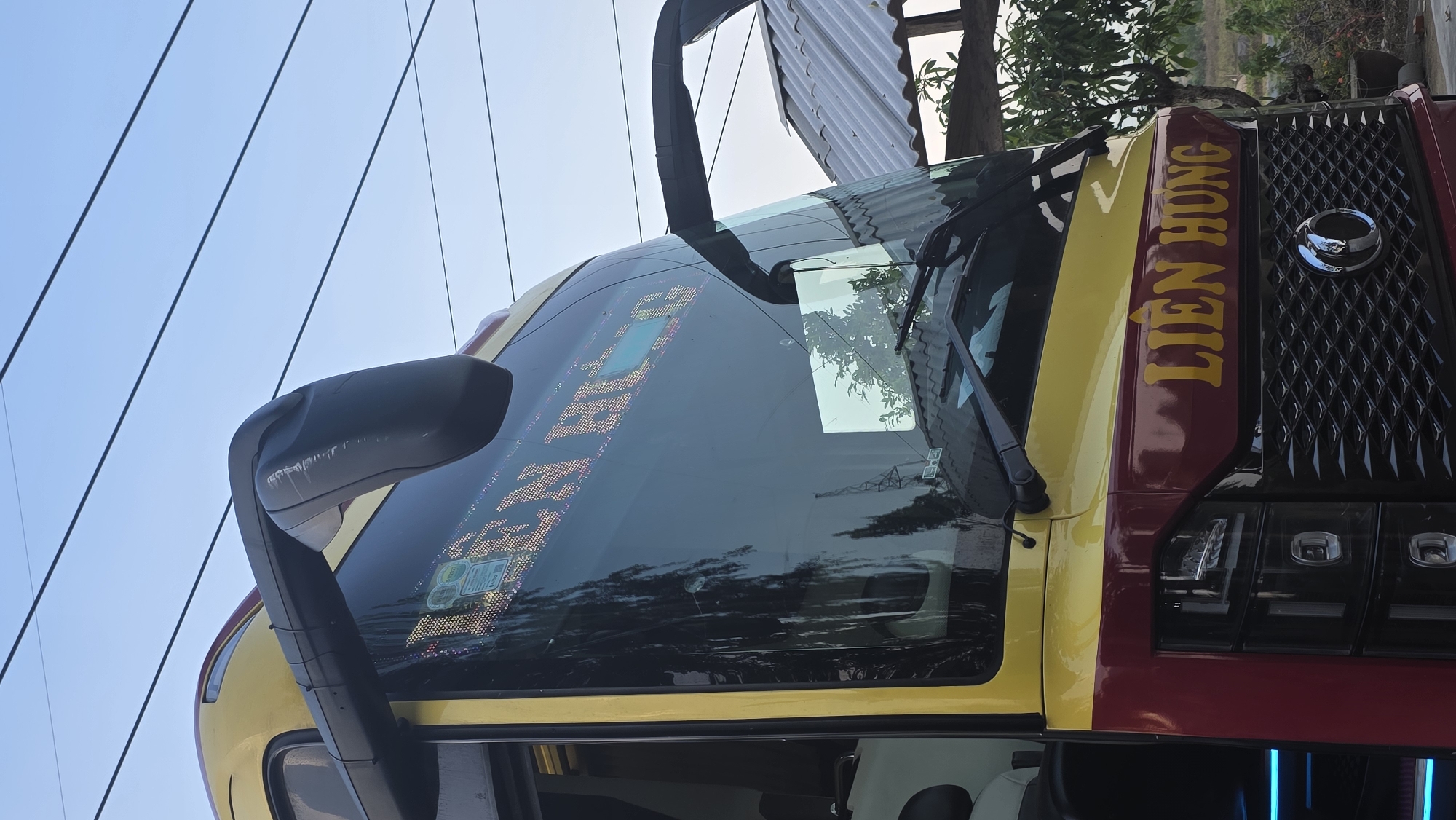A bus is photographed, its yellow-and-maroon paint scheme evident. The name "LIÊN HƯNG" is written in yellow on the vehicle's front. Power lines are visible against the sky, and vegetation is present in the background. The photo suggests a location in Ea Tê, Vietnam, based on the bus branding. The perspective is an upward shot, focusing on the front of the bus.